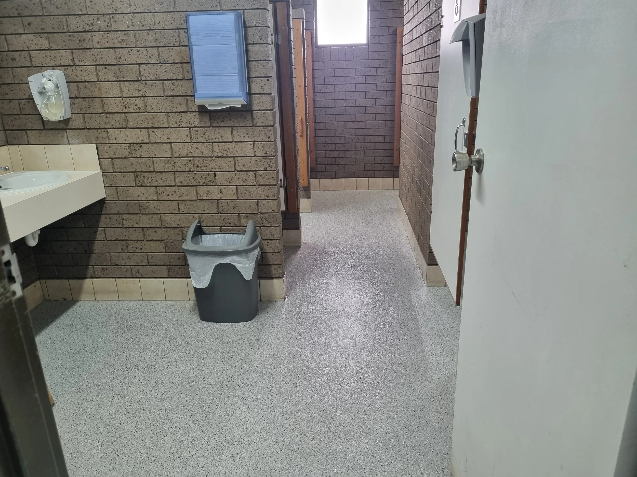 Public restroom with brick walls, a sink, a paper towel dispenser, a trash can, and stalls in the background.