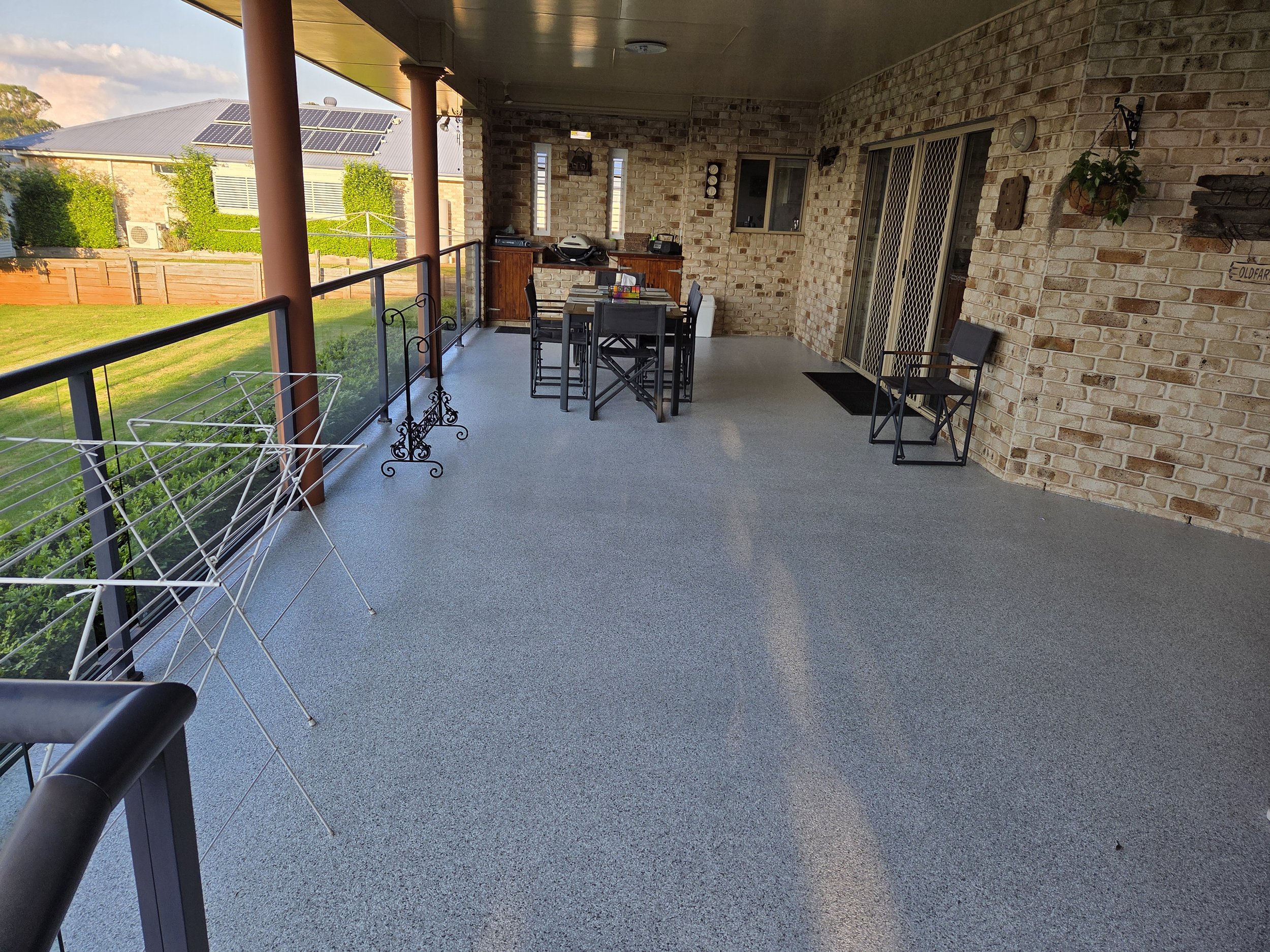 Covered outdoor patio with a brick wall, black metal chairs, a high table, clothing drying rack, and a barbecue grill, overlooking a backyard with grass and a wooden fence.