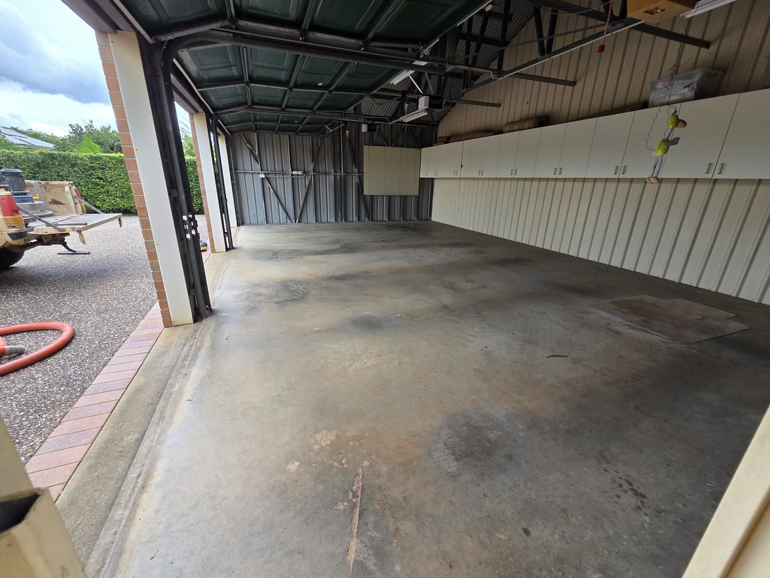 Empty garage with a concrete floor, metal walls, white cabinets along one wall, and an open garage door showing an outdoor area with a gravel driveway, a vehicle, and green hedges.