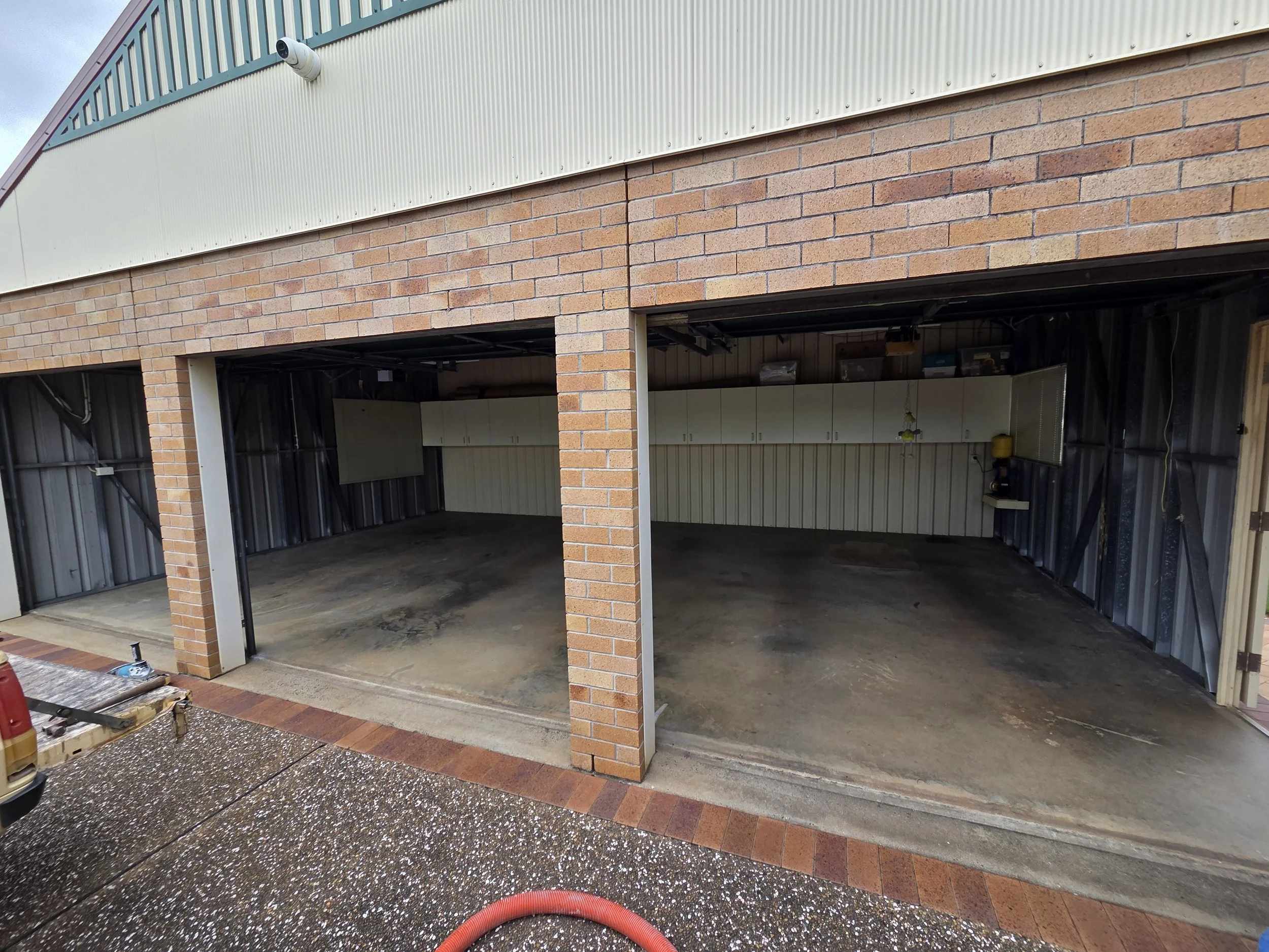 Two open garage parking spaces with brick and metal siding, empty concrete floors, and a small work area with cabinets and tools on the right wall.