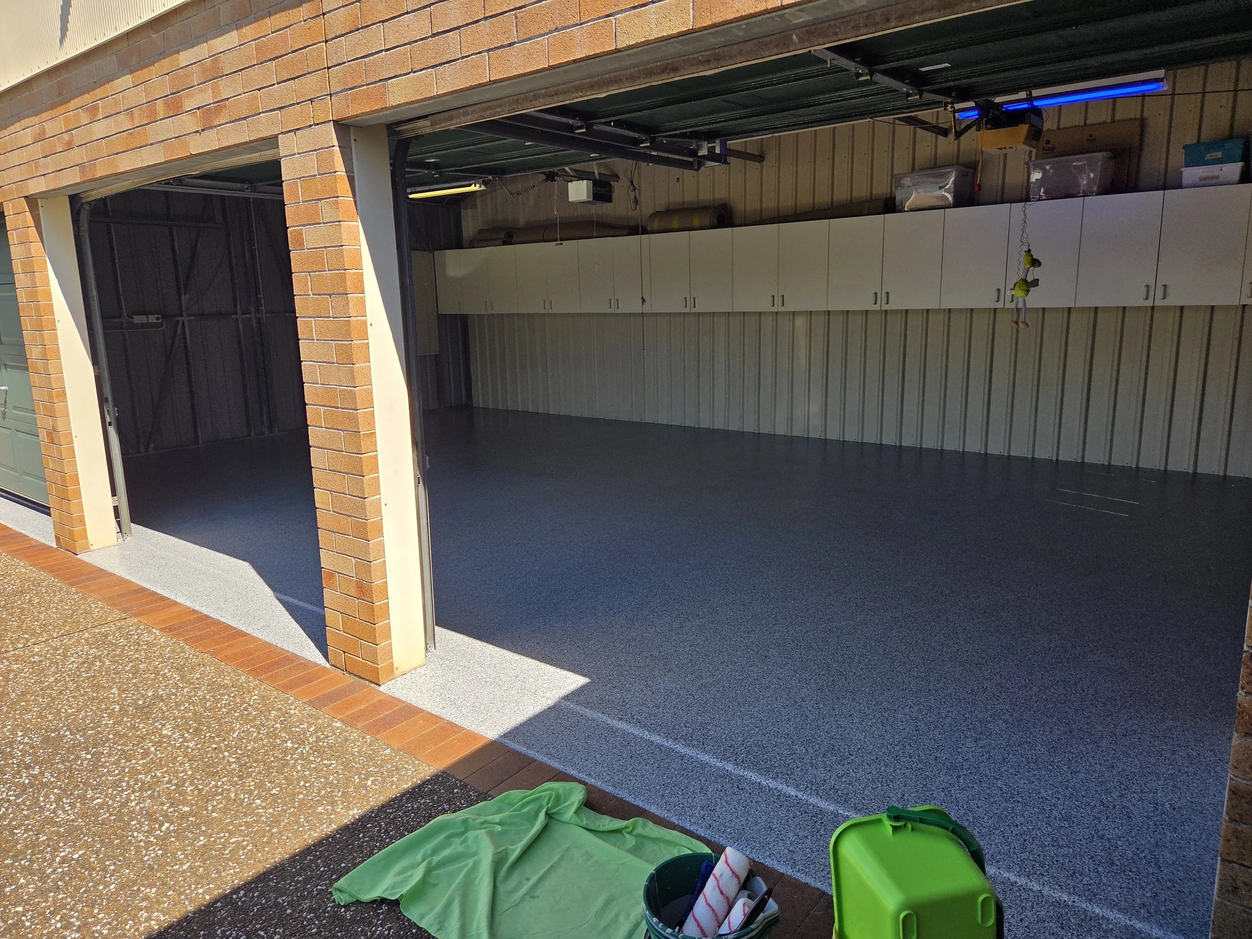 An open garage with newly painted gray flooring, white cabinets inside, and outdoor brick and concrete surfaces in sunlight.