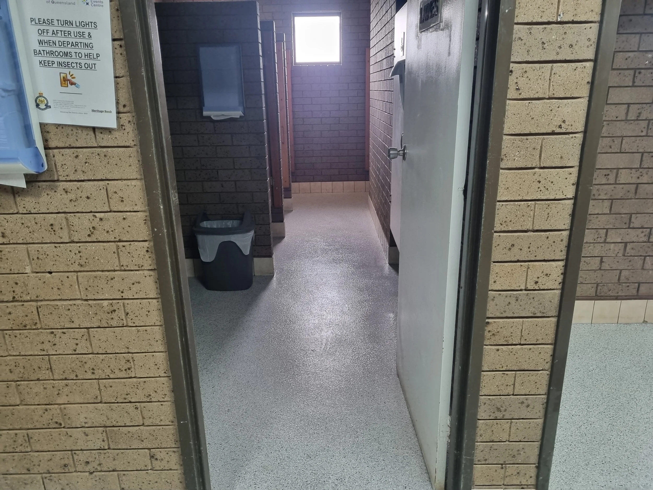 Public restroom with stall doors, a trash can near the stalls, brick walls, and a window providing natural light.