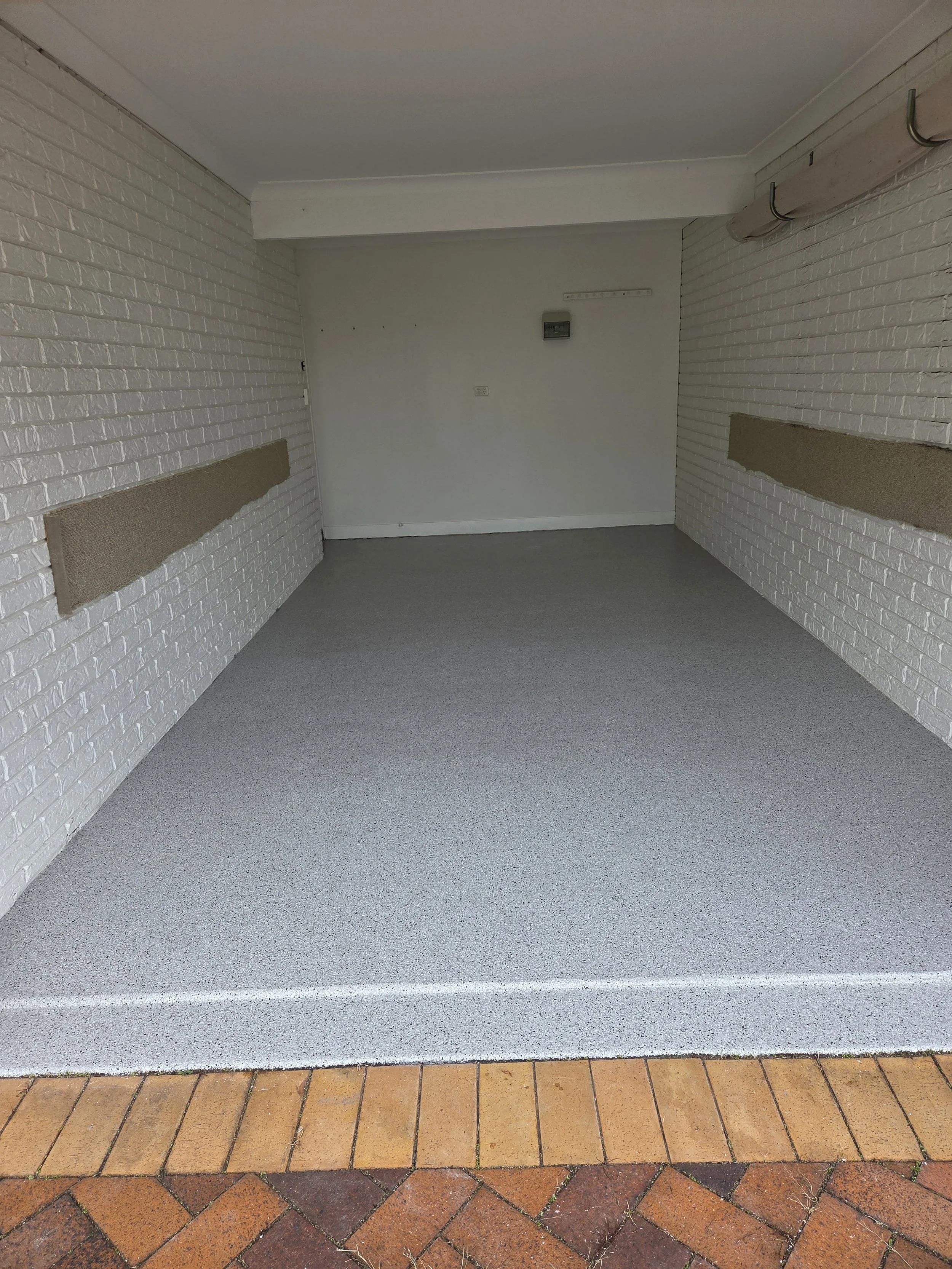 Empty garage with white brick walls, gray flooring, and a section of brick pavers at the entrance.
