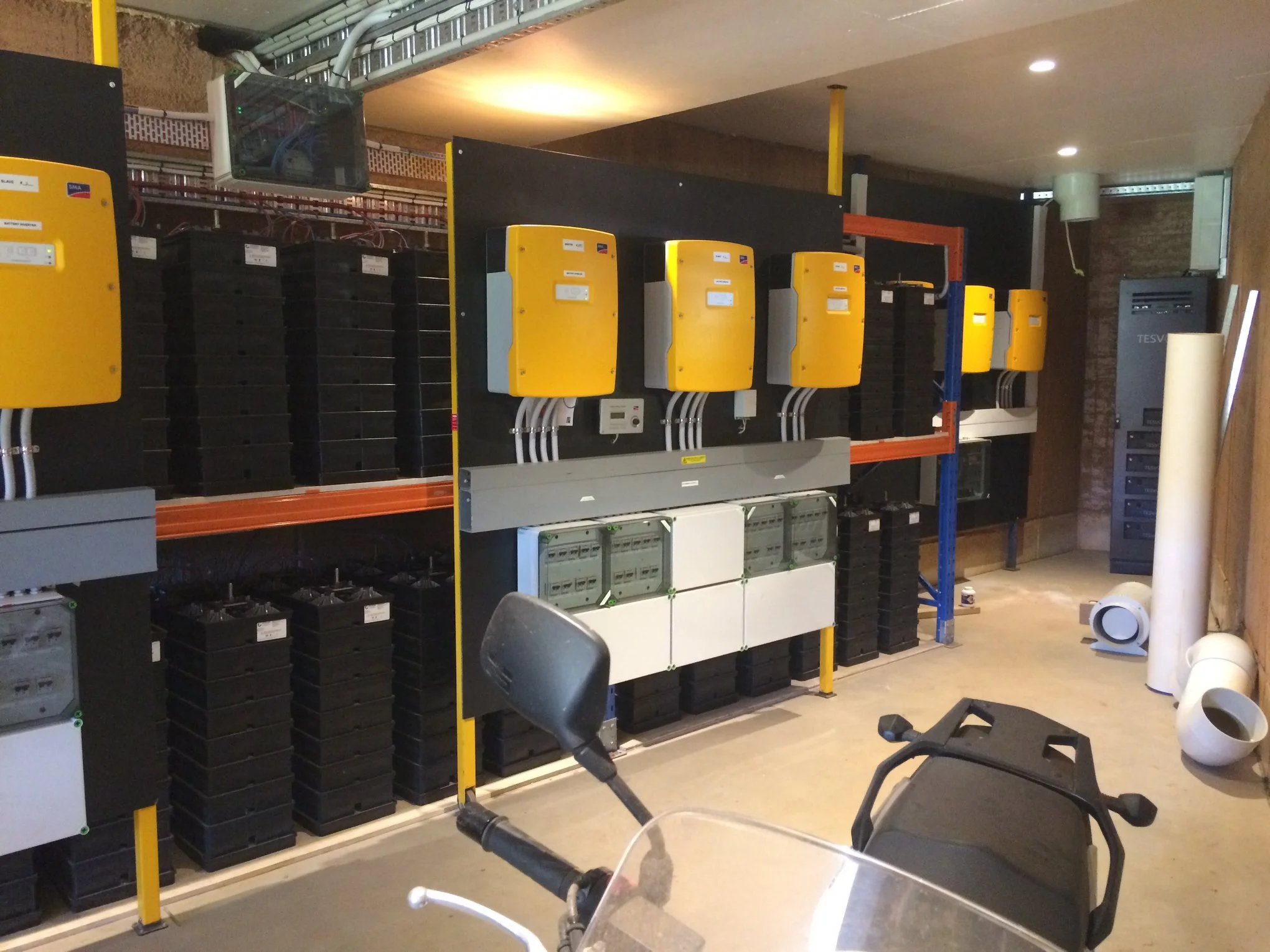 Electrical installation with yellow inverters, black batteries, and various electrical boxes on shelves, with some pipes and a motorcycle in the foreground.