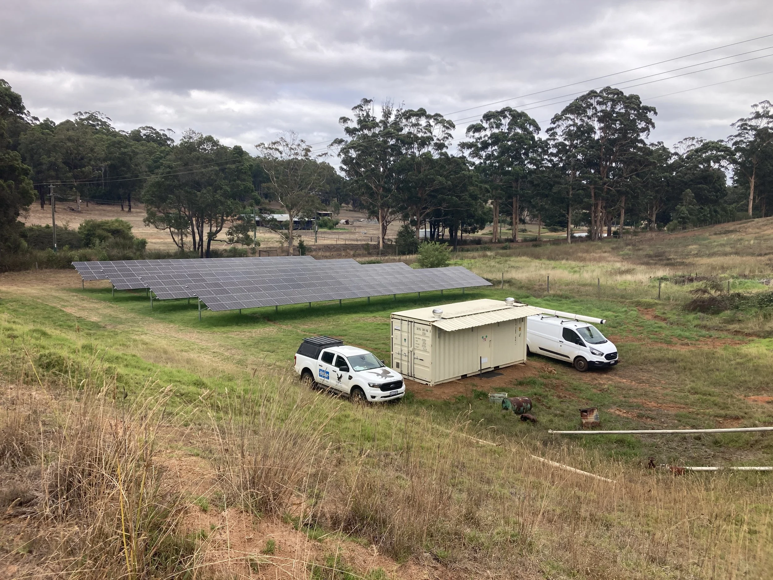 Solar panel farm with two white utility vehicles and a beige container on a grassy hill, surrounded by trees and overcast sky.