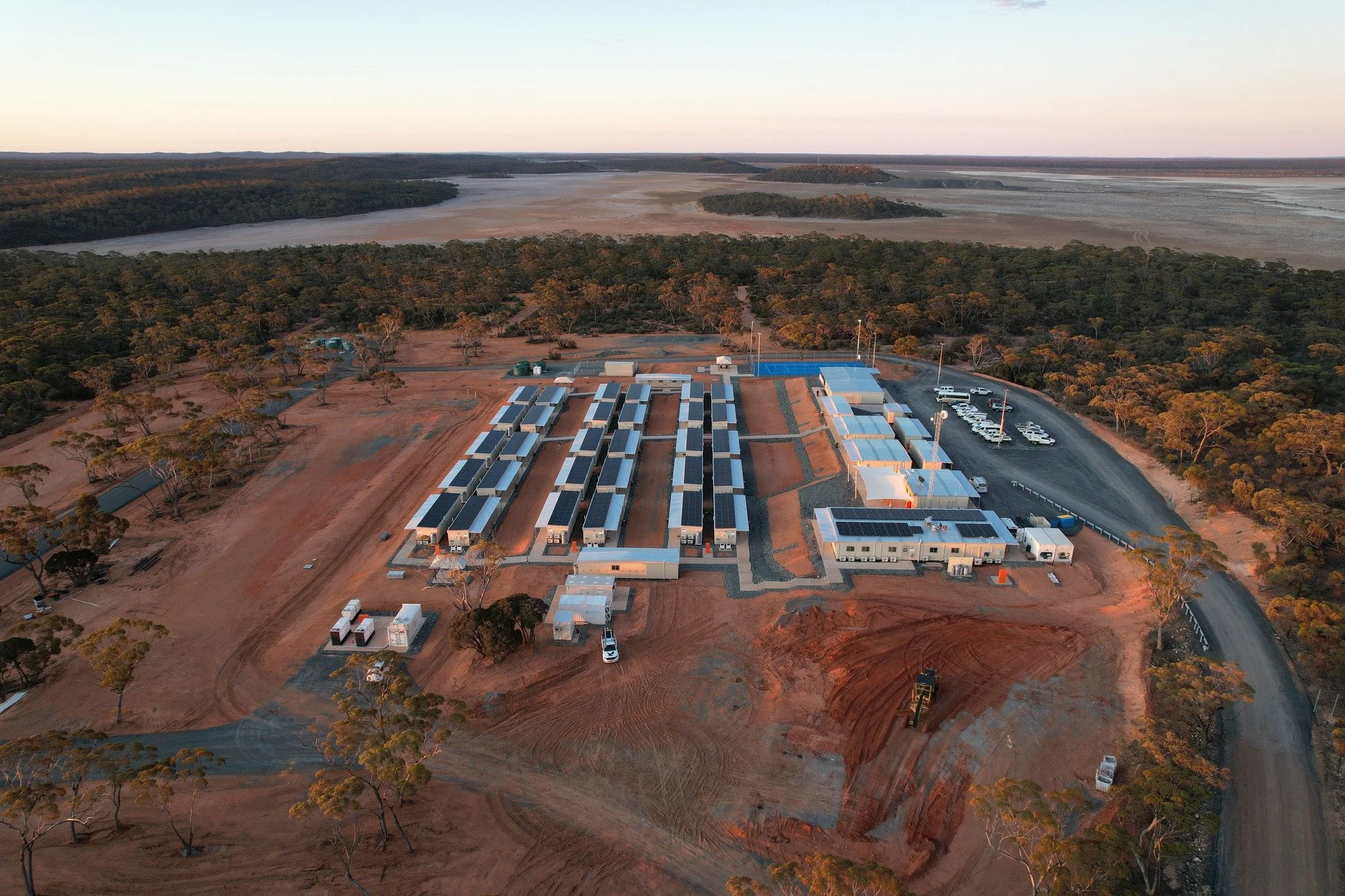 Aerial view of a solar power farm with multiple solar panel arrays on a cleared land, surrounded by trees and a nearby body of water.