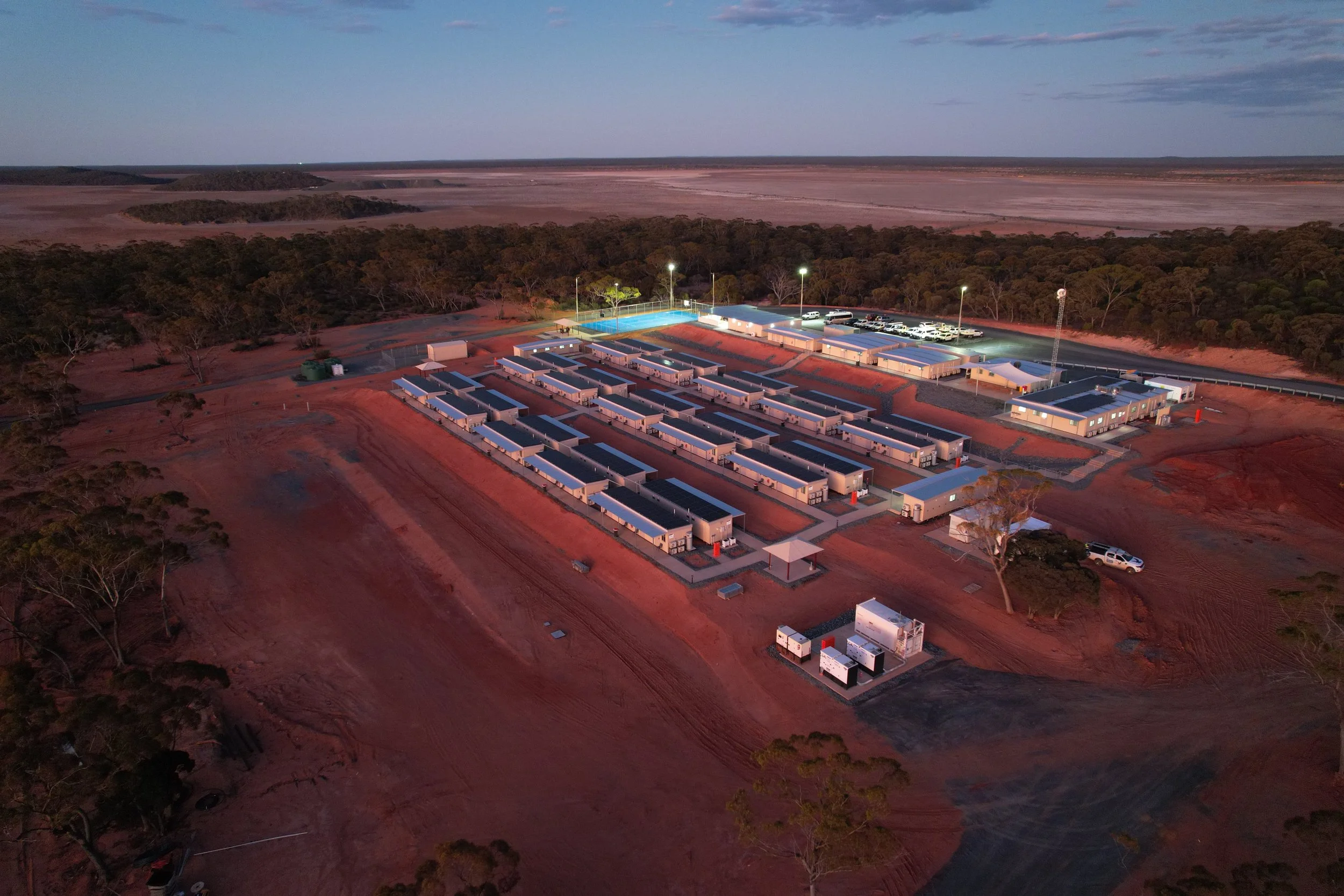 Aerial view of a large, illuminated outdoor sports and recreation complex in a remote area, surrounded by red dirt and sparse trees, with a distant horizon and open sky.