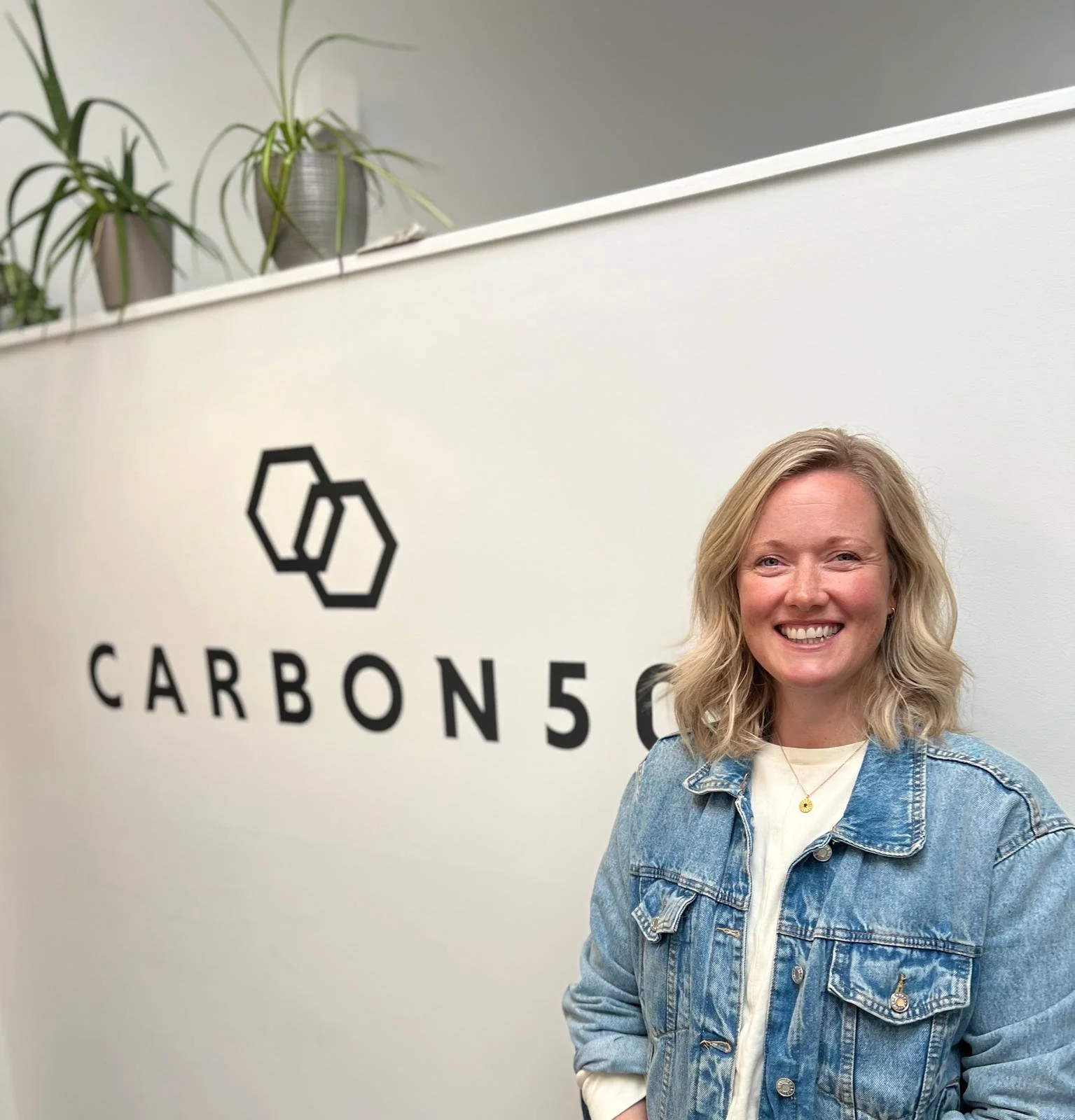 A smiling woman with blonde hair wearing a denim jacket standing in front of a wall with the Carbon5 logo and plants on a shelf.