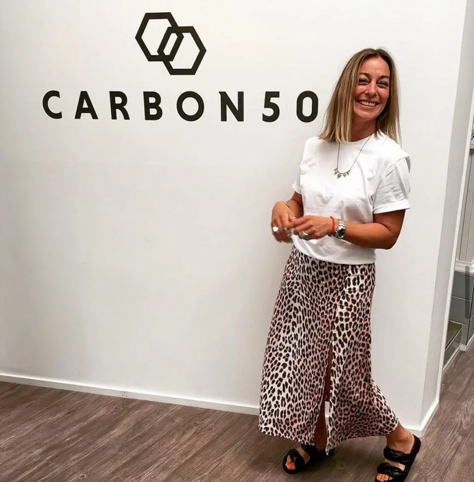 A woman standing in front of a white wall with a logo and the text "CARBON50," smiling and wearing a white t-shirt, leopard print skirt, and black sandals.