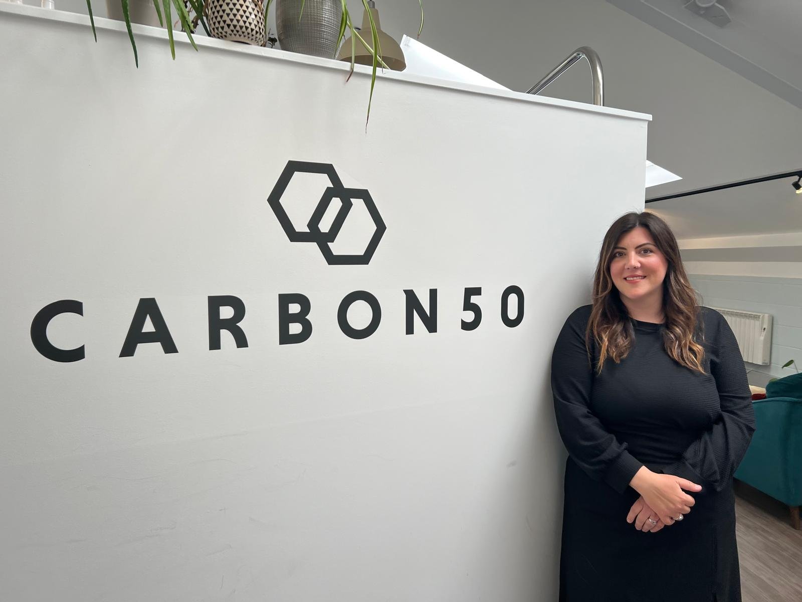 A woman with long brown hair wearing a black dress standing next to a white wall with the logo and text 'CARBON50' and a hexagon design, inside a modern office or conference space.