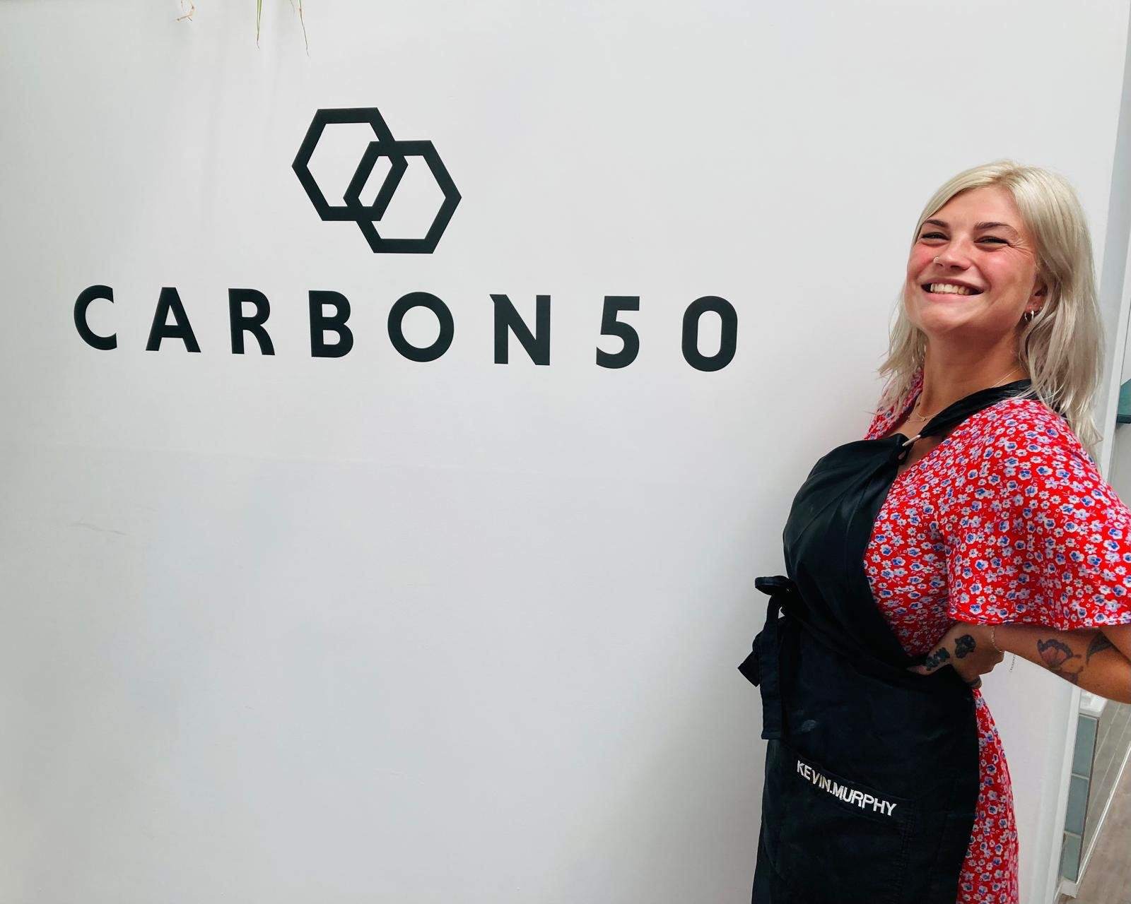A woman with blond hair, smiling, standing in front of a white wall with a logo and text that reads 'CARBON50'. She is wearing a red floral dress and a black apron with the name 'Kevin Murphy' on it.