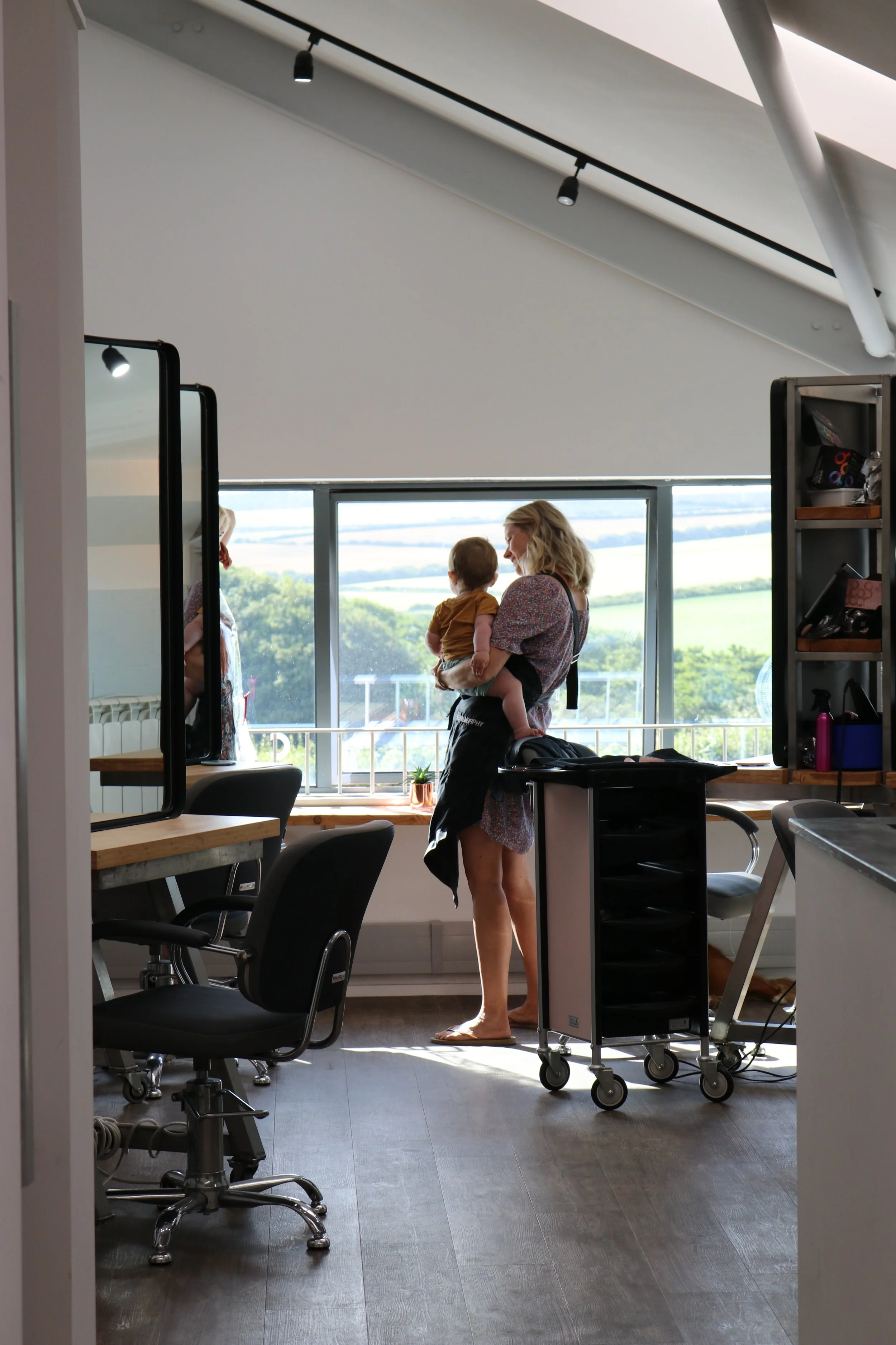 Woman holding a child and looking out a large window in a modern room with office chairs, a mirror, and open shelves.