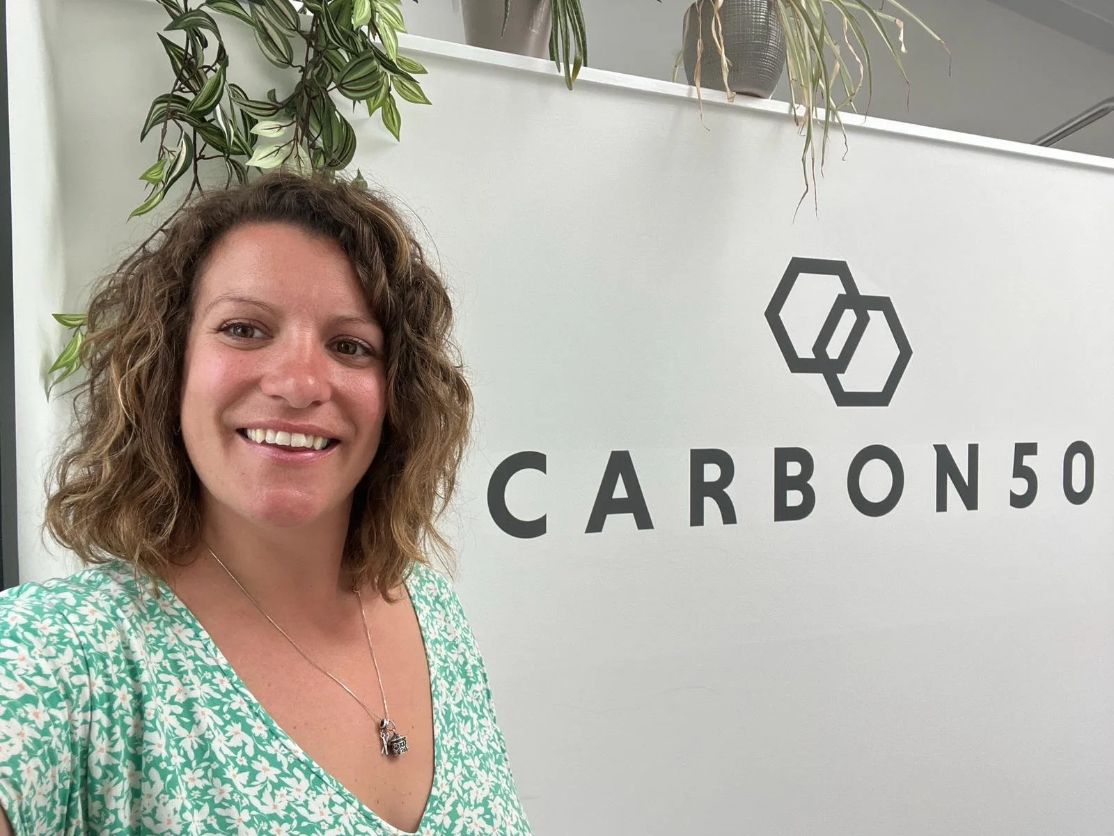 A woman with curly brown hair smiling in front of a white wall with 'CARBON50' and a logo of two interlinked hexagons. Potted plants are on top of the wall.