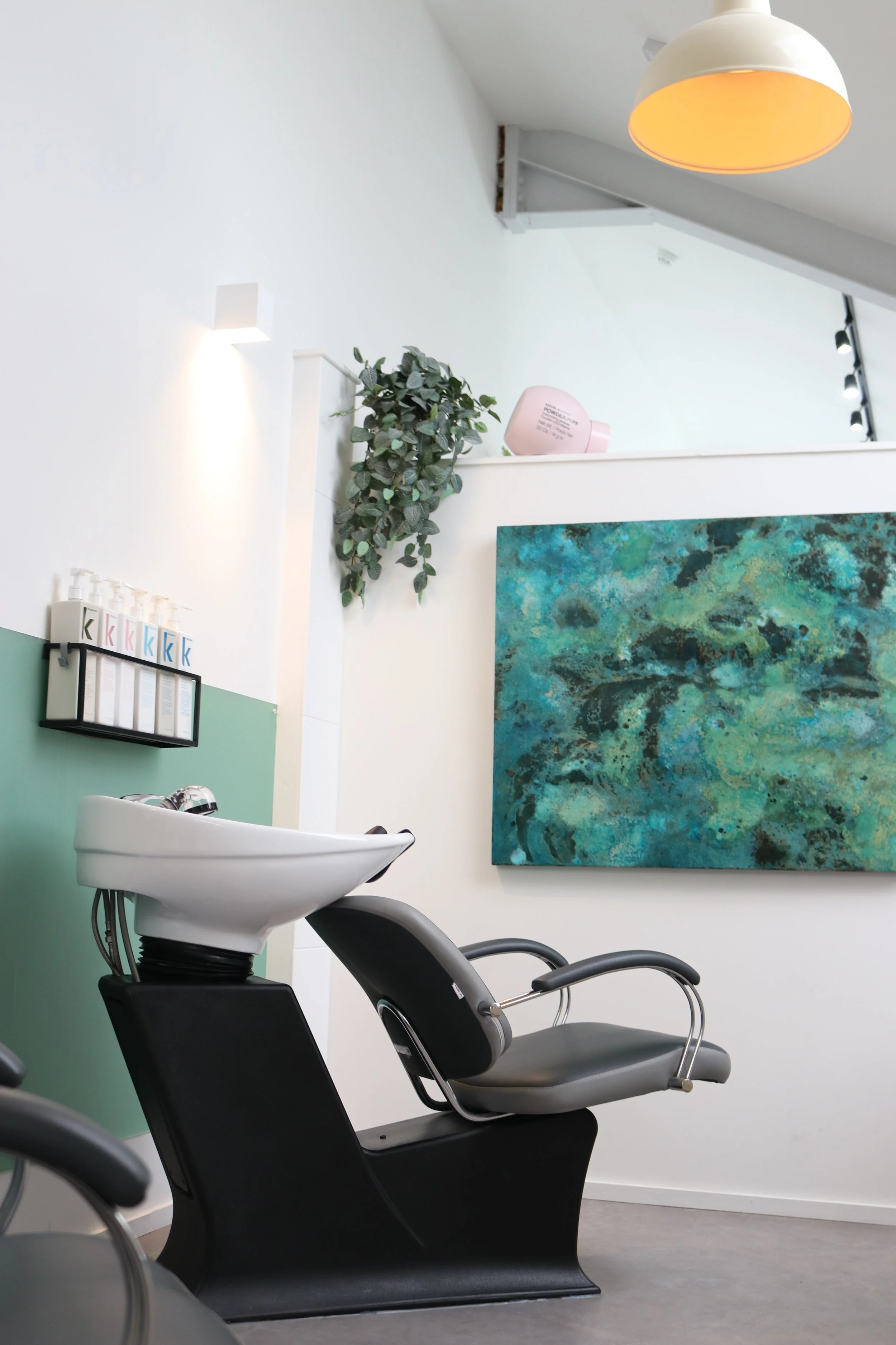 An empty hair salon shampoo station with a white sink, black chair, and green wall panel. There is abstract blue and green artwork on the white wall. A small shelf with hair products, a hanging plant, and a pink device are visible.