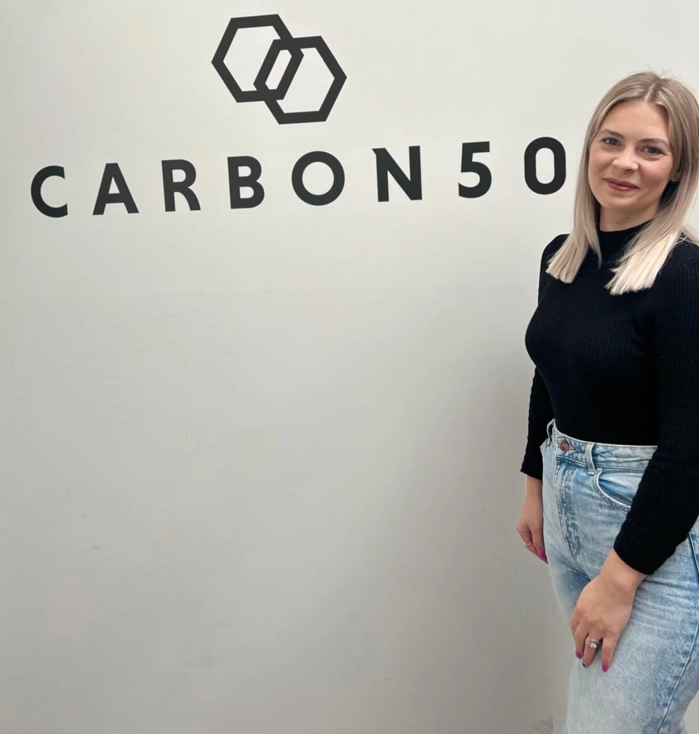 A woman with blonde hair wearing a black sweater and light blue jeans standing next to a wall with a logo and the text 'CARBON 50' on it