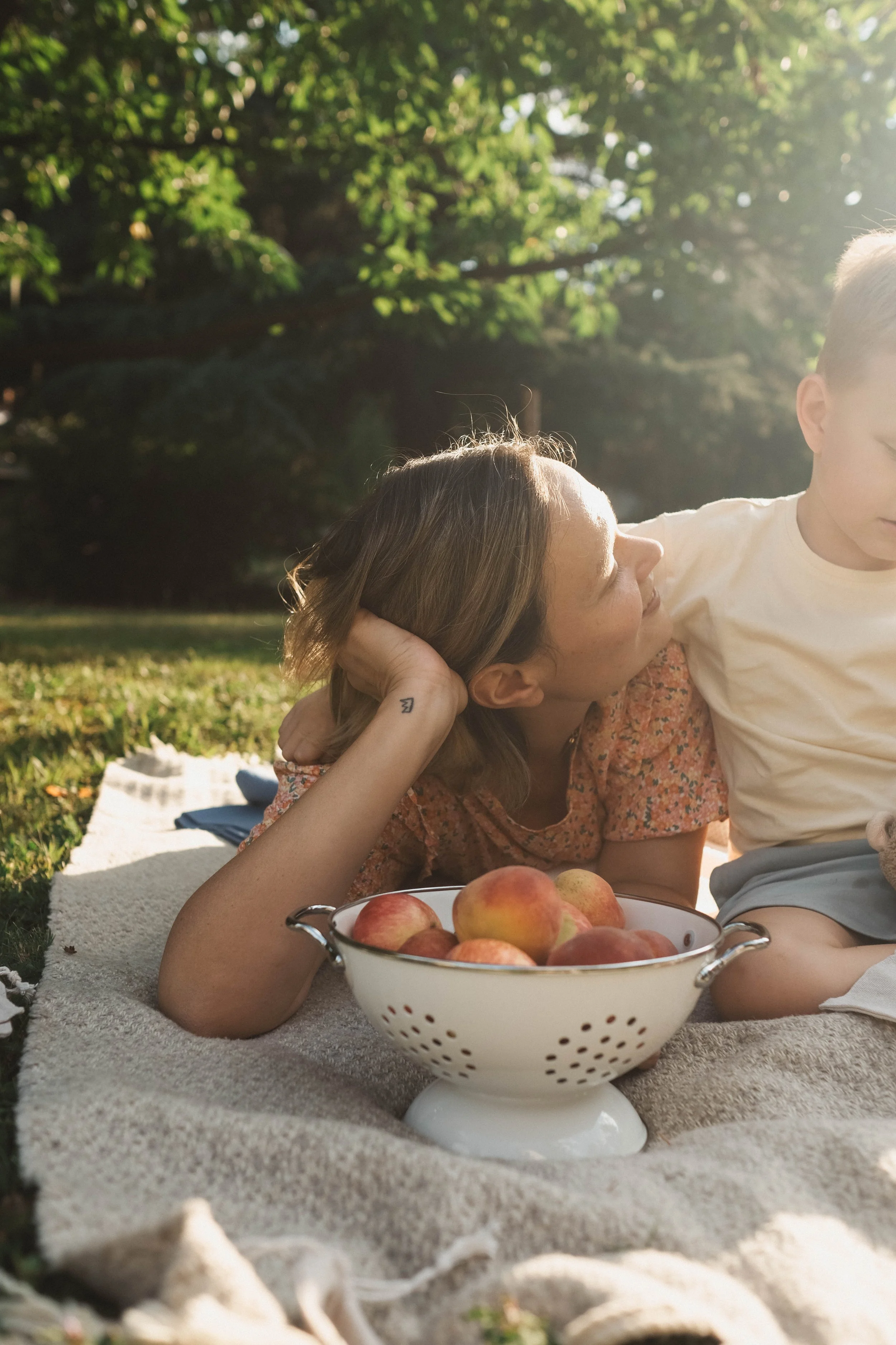 A woman and young boy lying on a blanket outdoors during sunlight. The woman is looking at the boy while resting her head on her hand. There is a bowl of peaches next to them on the blanket.