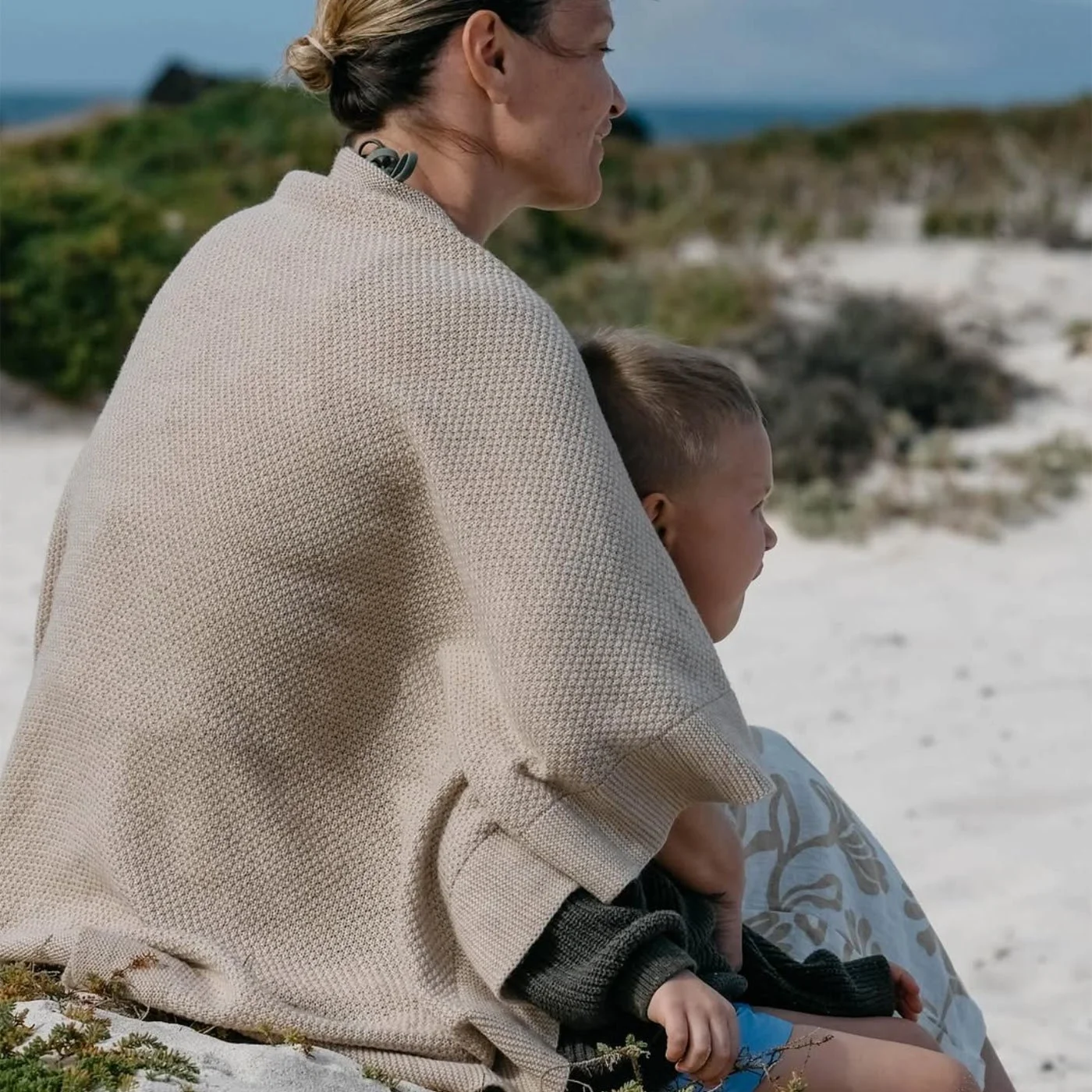 A woman with a young child sitting on the sandy beach, facing the ocean, with dunes and green shrubs in the background.