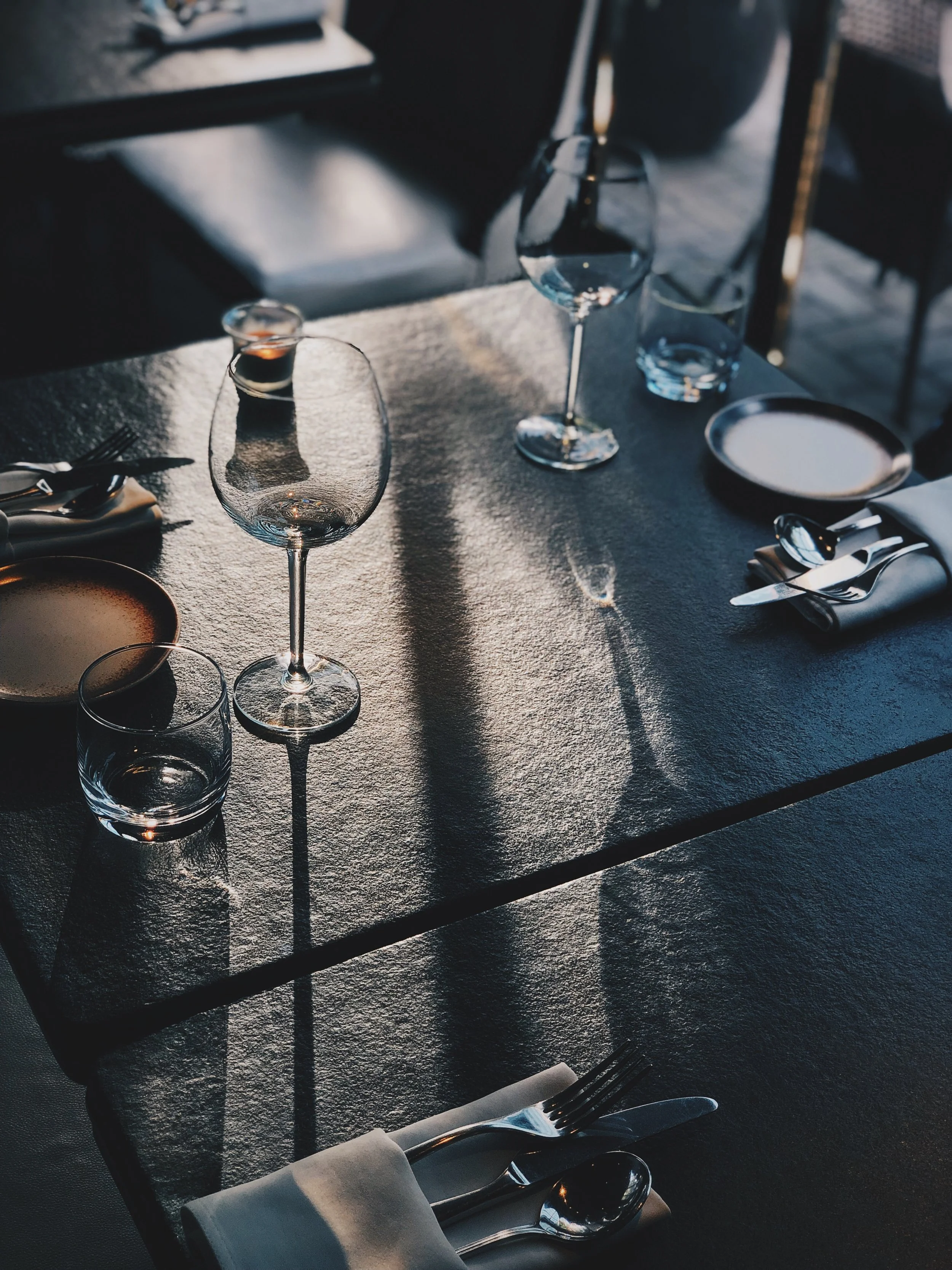 Set dining table with wine glasses, water glasses, plates, silverware, and a folded napkin, with dramatic lighting creating shadows and reflections.