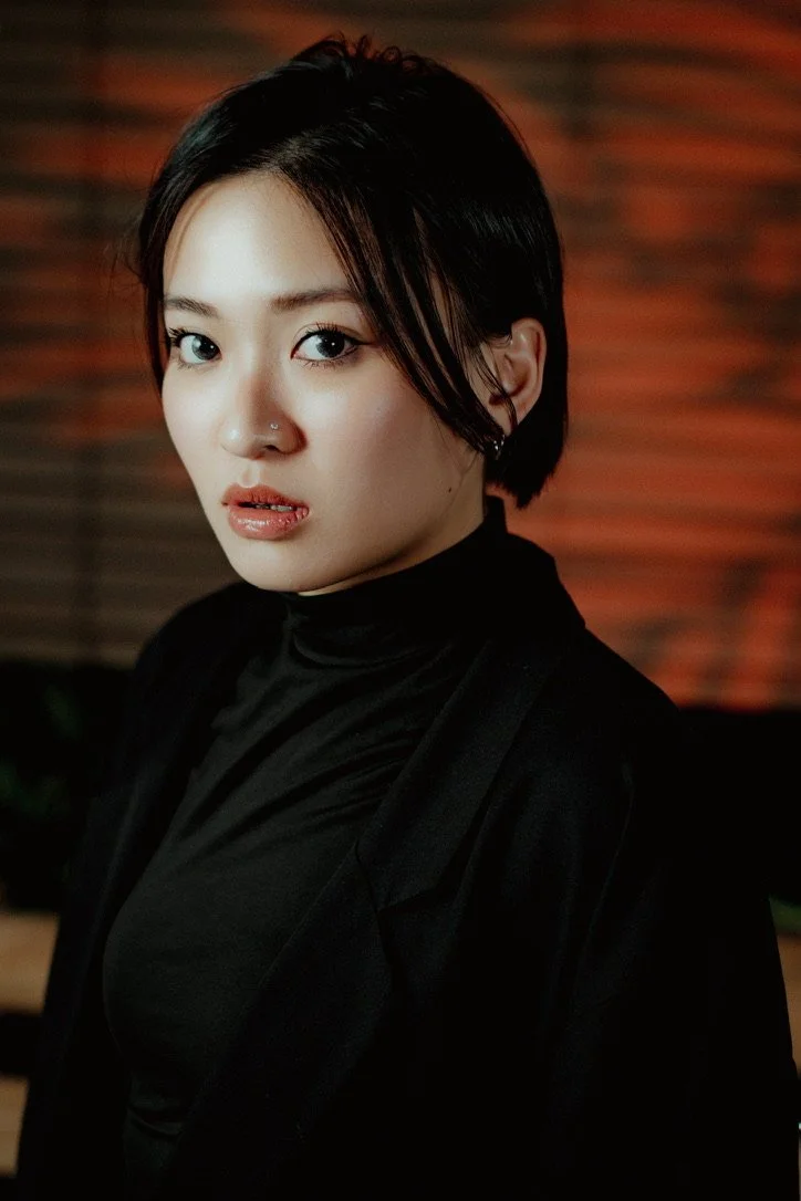 A woman with short black hair, blue eyes, and a nose piercing looks at the camera against a blurred brick wall background.