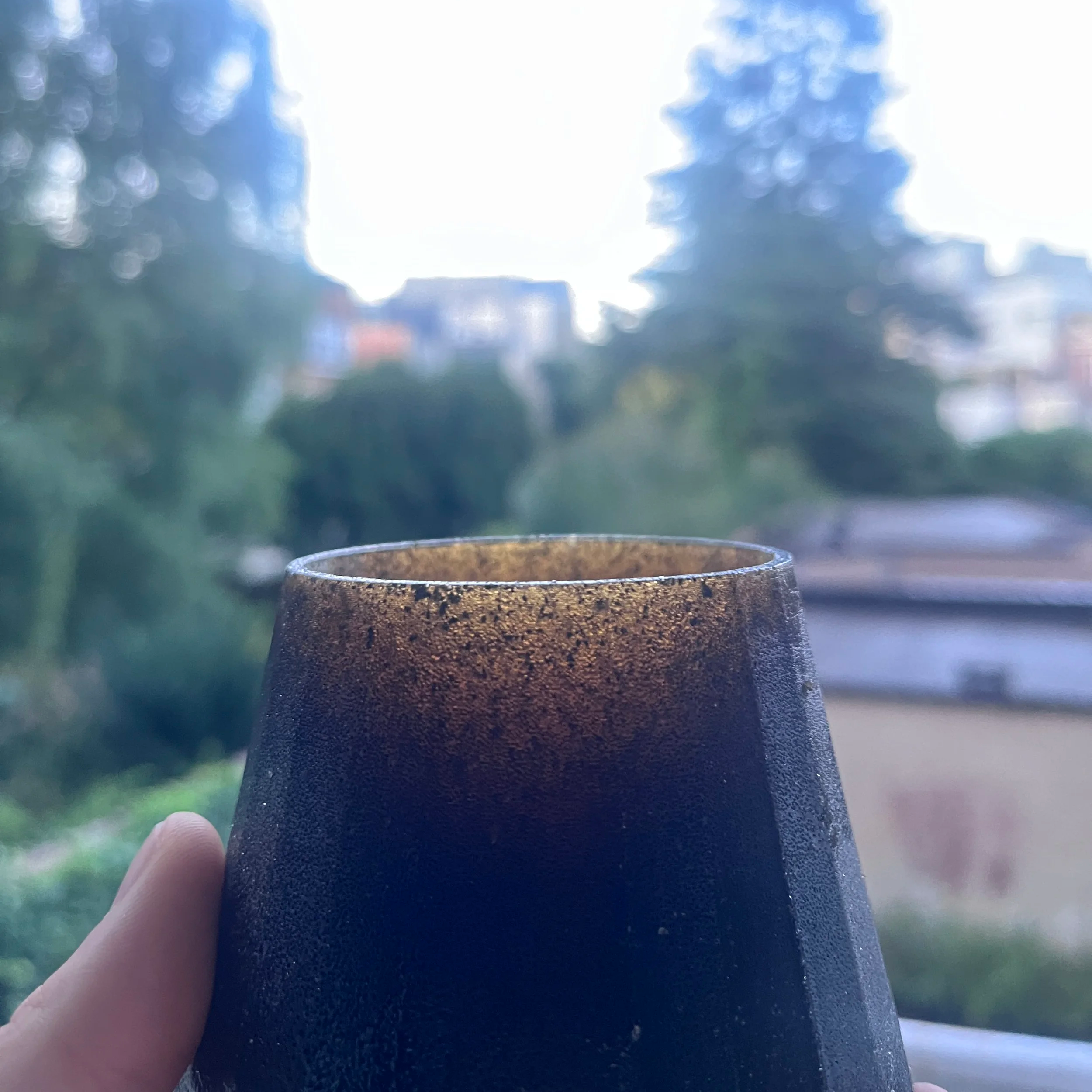 Una mano tiene una tazza di vetro con un bordo dorato. Lo sfondo mostra un paesaggio con alberi e case sfocate, alla luce del sole.