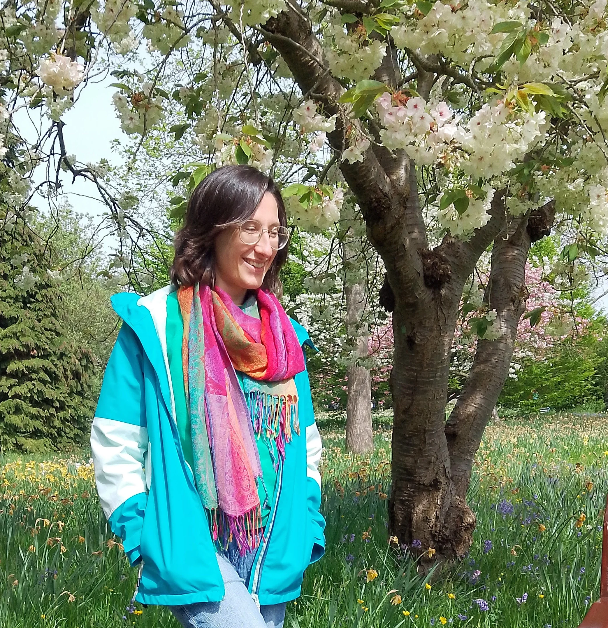 A woman with glasses smiling standing outdoors under flowering trees, wearing a colorful scarf and a blue jacket.