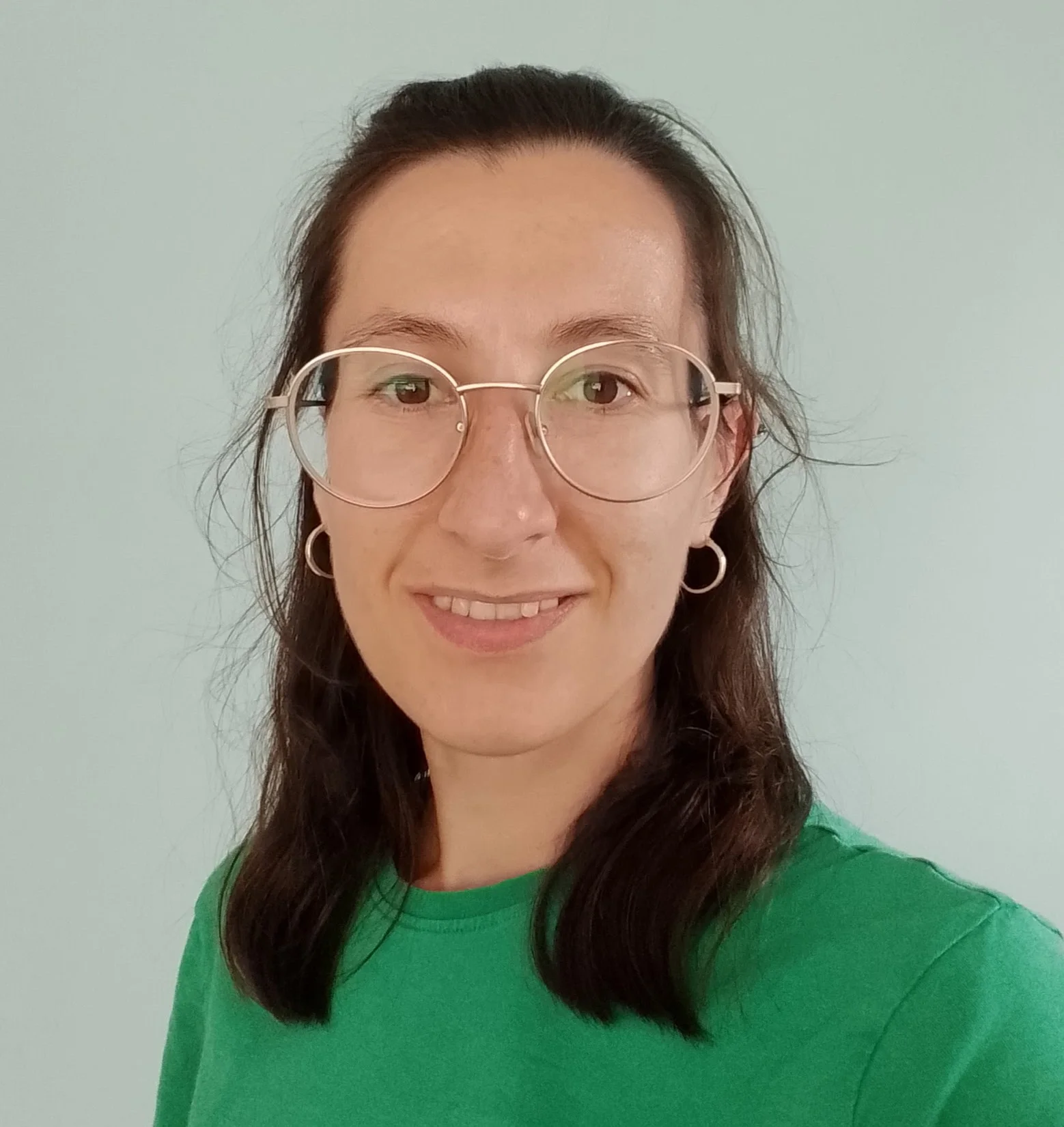 A woman with glasses and earrings smiling in front of a plain light-colored wall, wearing a green shirt.