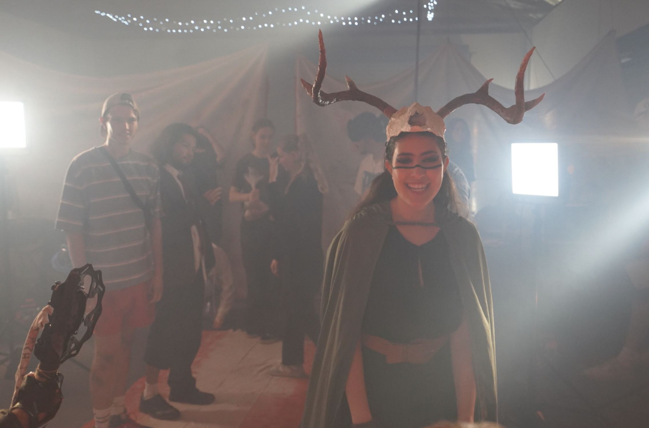 A woman dressed in a costume with antlers, face paint, and a cape, smiling at the camera at what appears to be a costume party or event. There are several people in the background, some engaged in conversation, with bright lights and a festive setting.