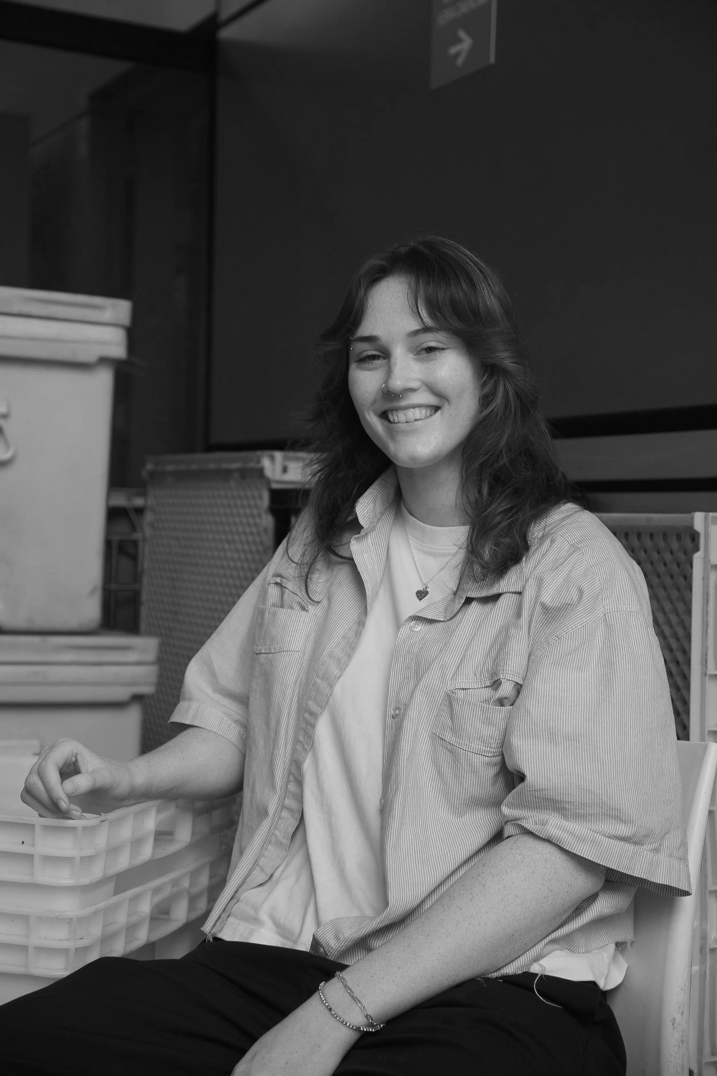 A young woman with shoulder-length wavy hair and a nose piercing, smiling while sitting next to plastic containers in an indoor setting.