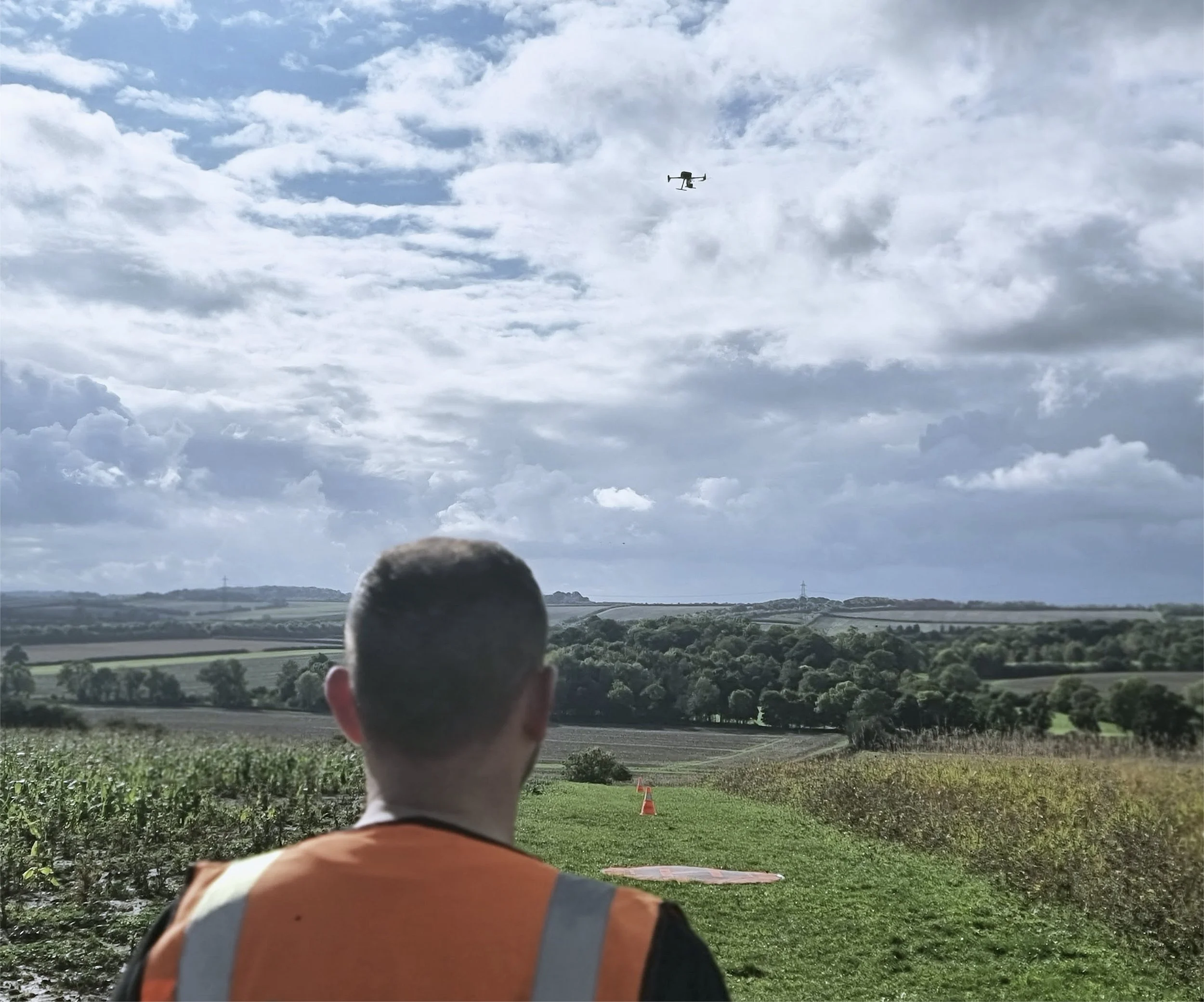 A person wearing an orange vest looks at a drone flying in a partly cloudy sky over a rural landscape with fields and trees.