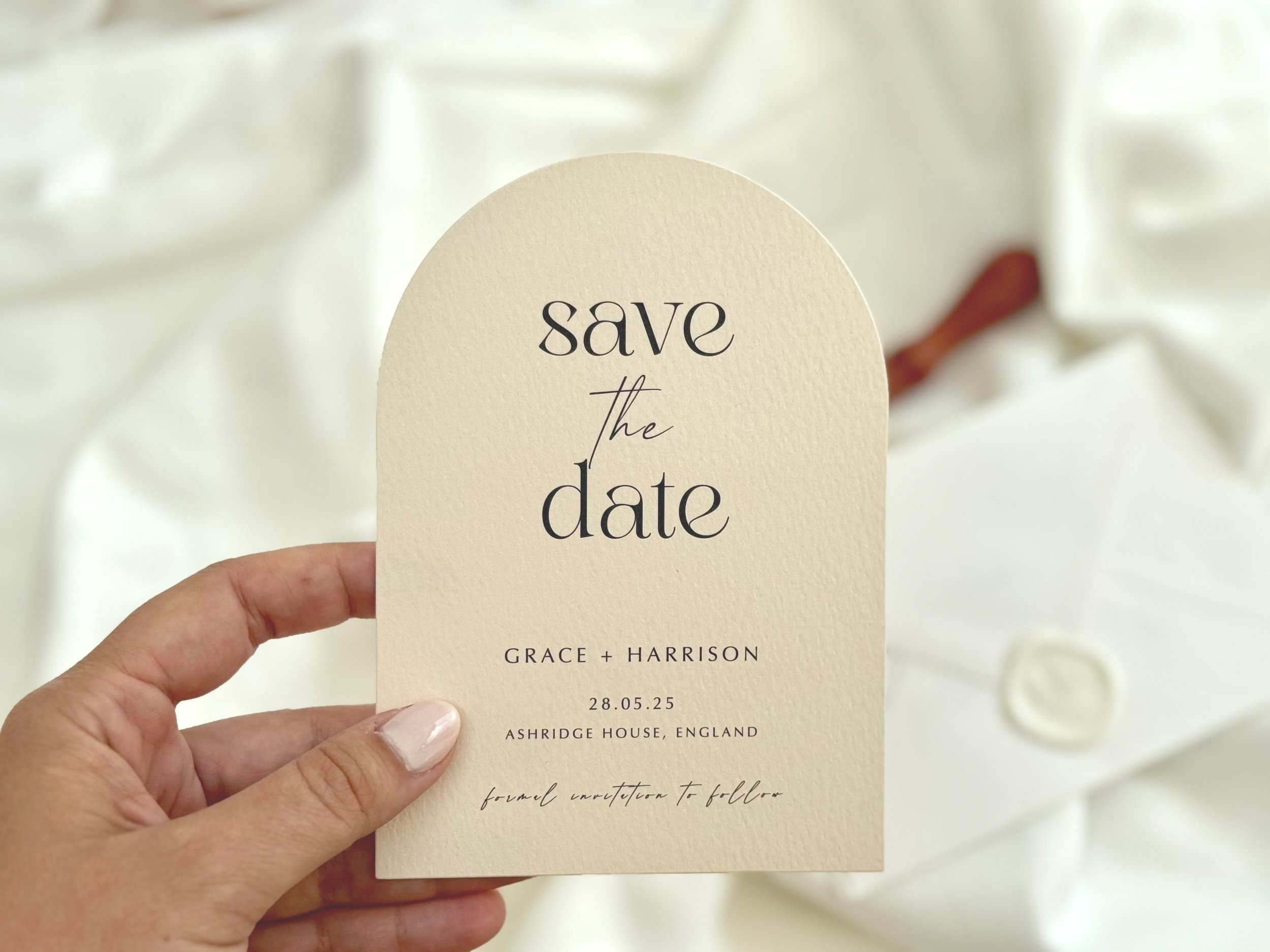 A hand holding a cream-colored save the date card with black and cursive writing for a wedding, mentioning Grace and Harrison, with details of the wedding date and venue, against a background of white fabric and a wrapped box.