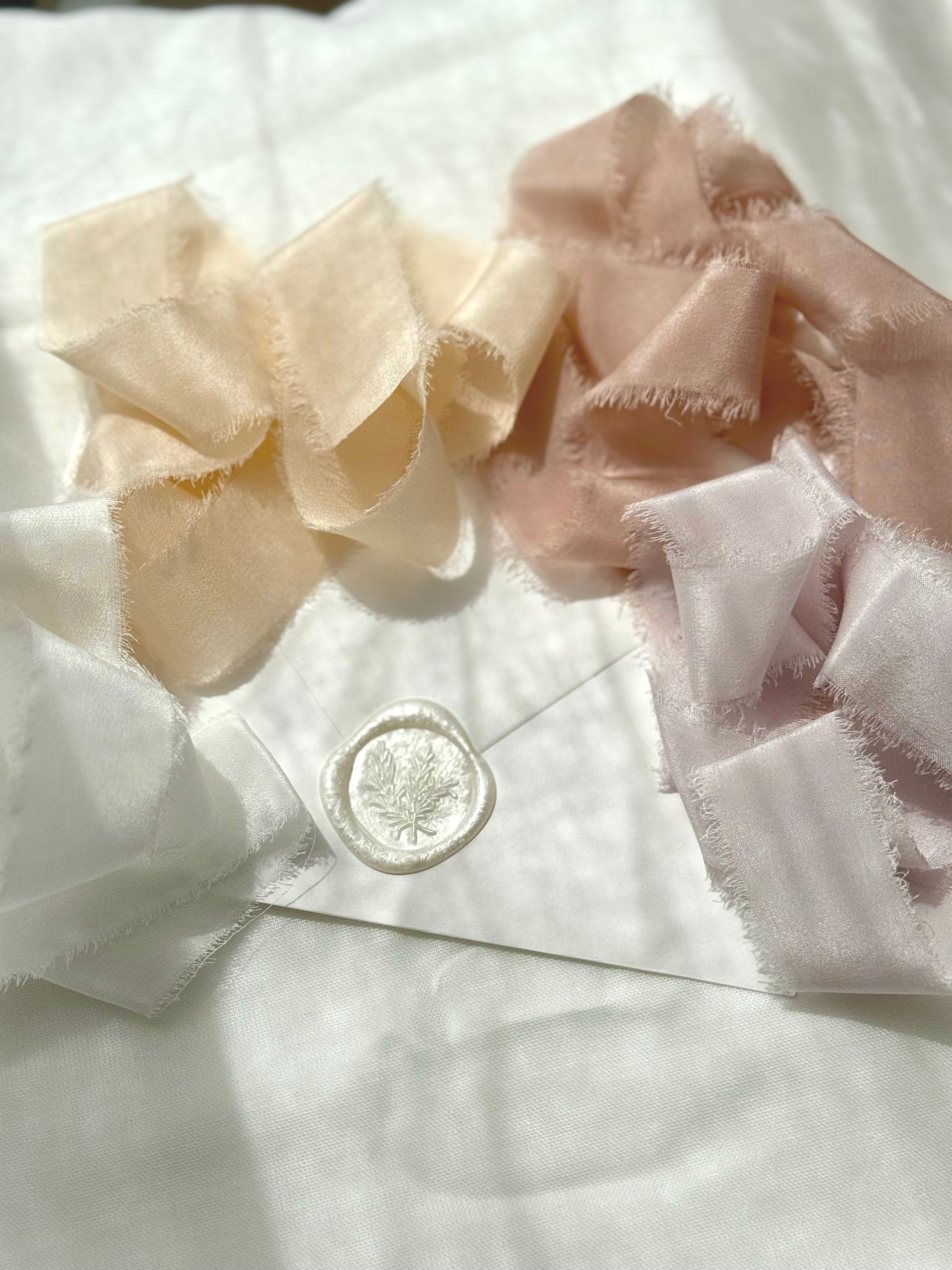Three fabric scrunchies in cream, pink, and white colors laid on a white fabric surface with a sealing wax stamp with a leaf design placed underneath them.
