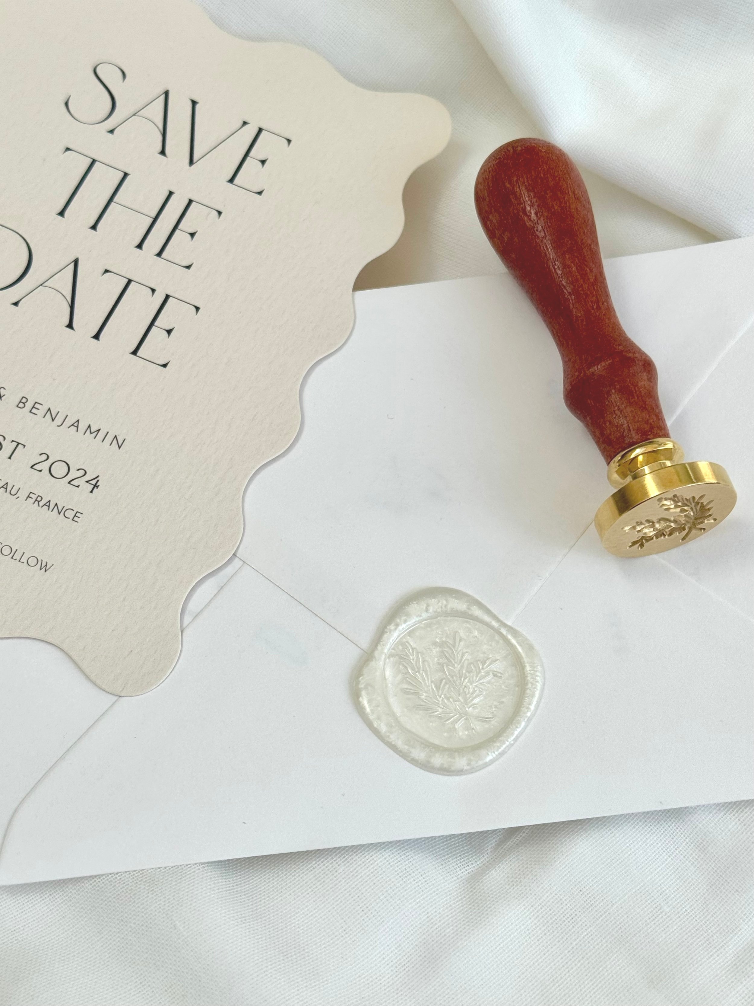 Save the date card with a wax seal, a stamp with a tree emblem, and a white envelope on a white surface.