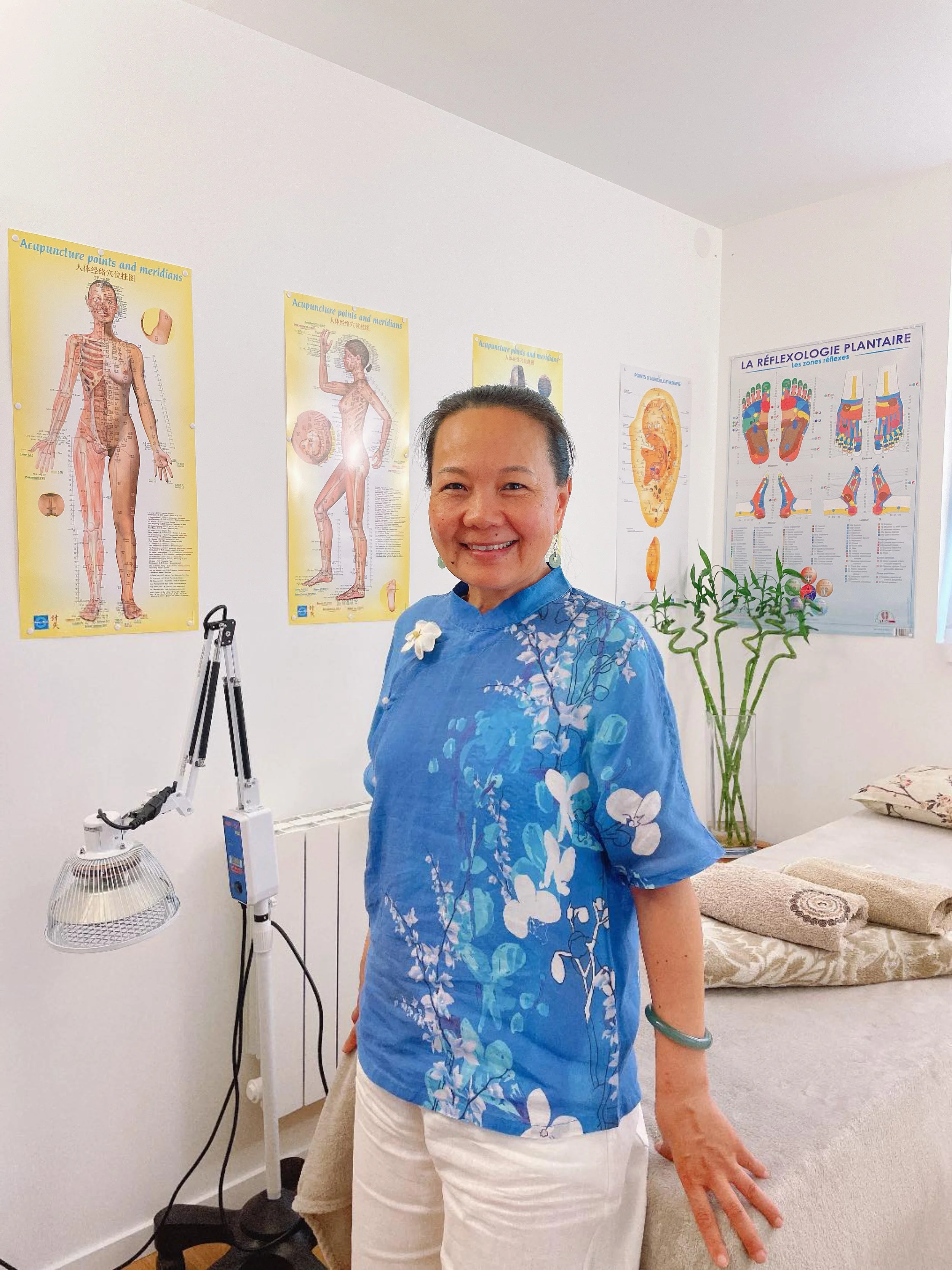 Une femme souriante portant une blouse bleue avec un motif floral dans une salle de thérapie, avec des posters de réflexologie plantaire et d'acupuncture sur les murs.