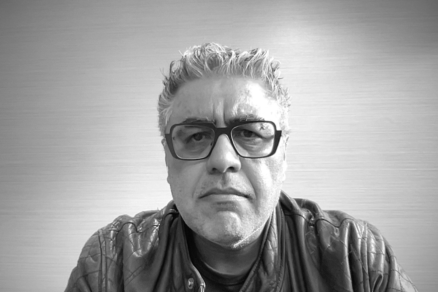 Black-and-white photo of a middle-aged man with curly hair and glasses, wearing a leather jacket, looking serious.