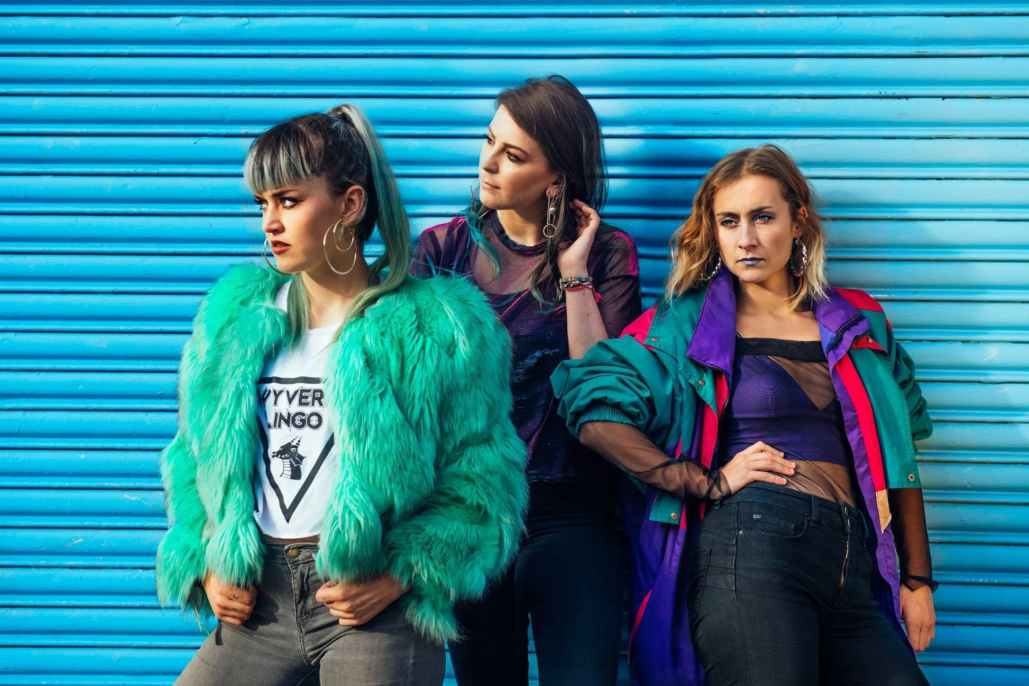 Three women standing in front of a blue corrugated metal wall, wearing colorful and edgy outfits with a retro vibe.