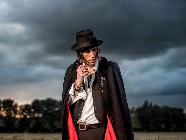 A person dressed as Michael Jackson wearing a black fedora, black jacket, beige shirt, and red scarf, standing outdoors under dark, cloudy skies.