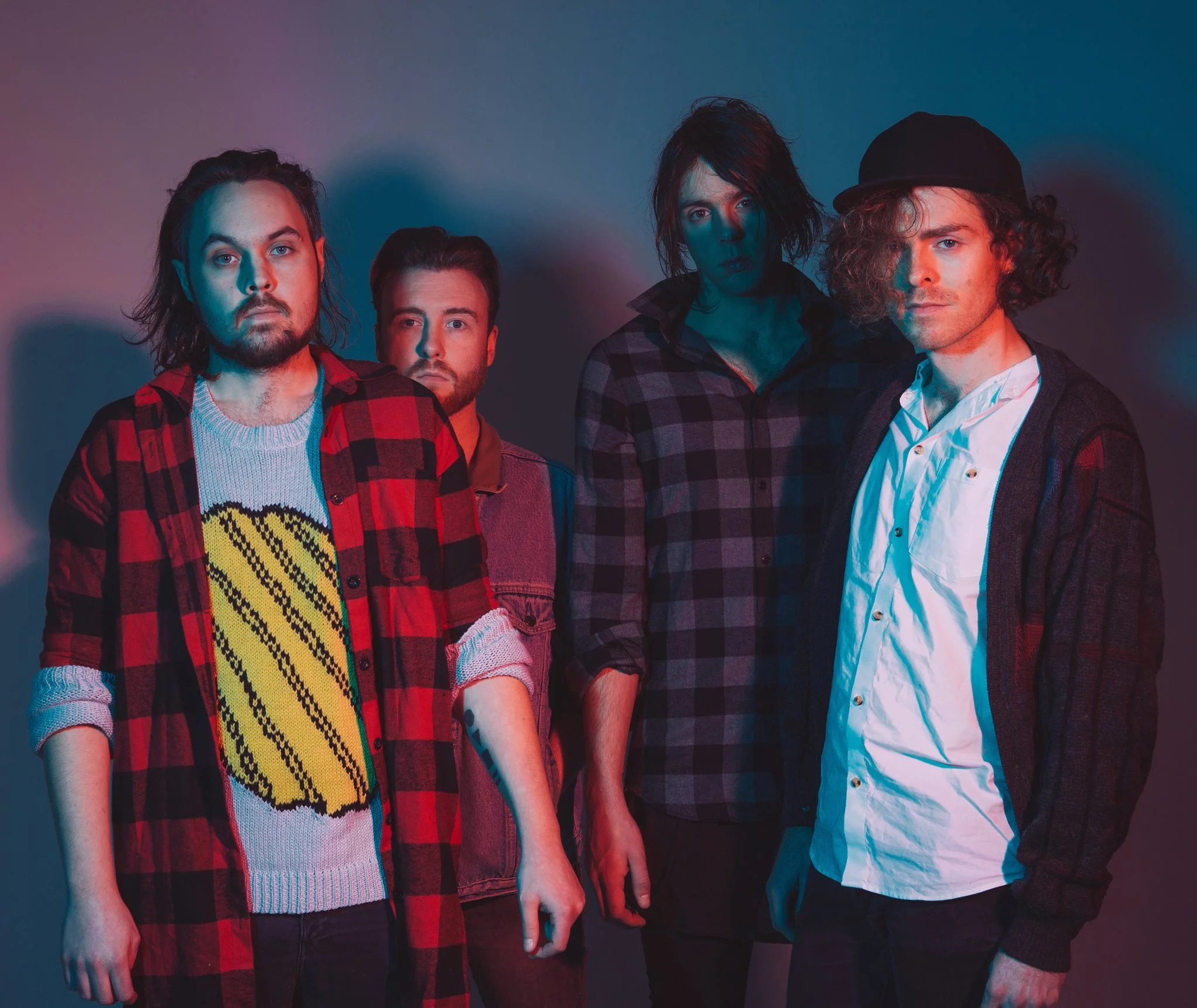 A group of four men standing against a wall with colorful lighting, dressed in casual and plaid clothing, with serious expressions.