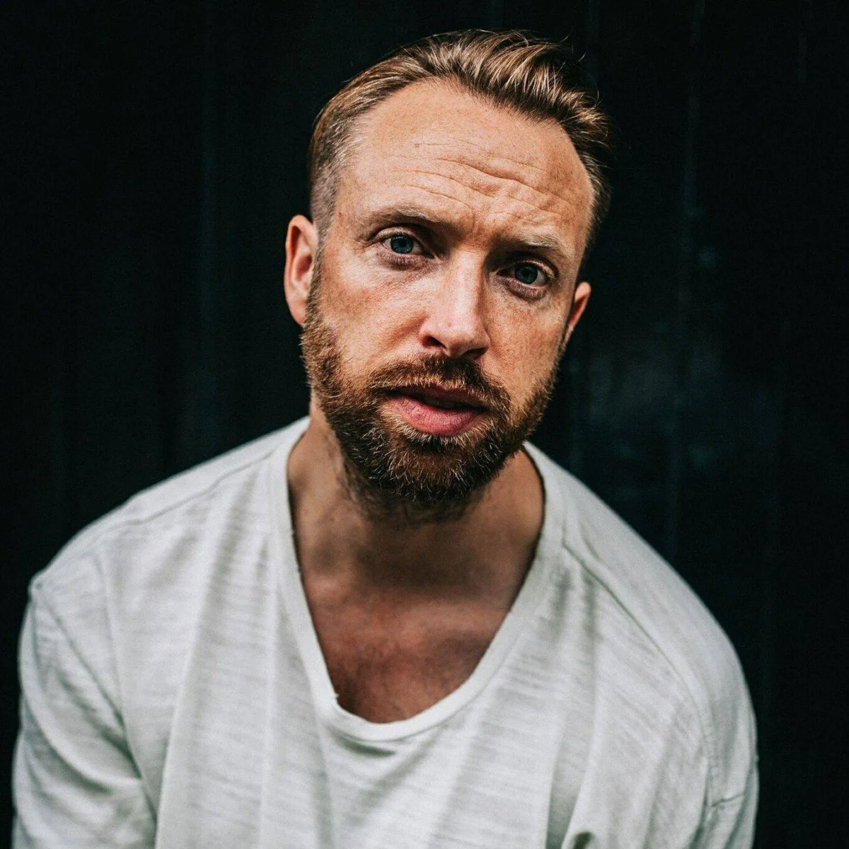 A close-up portrait of a man with light skin, blue eyes, short light brown hair, and a beard, wearing a white T-shirt, against a dark background.