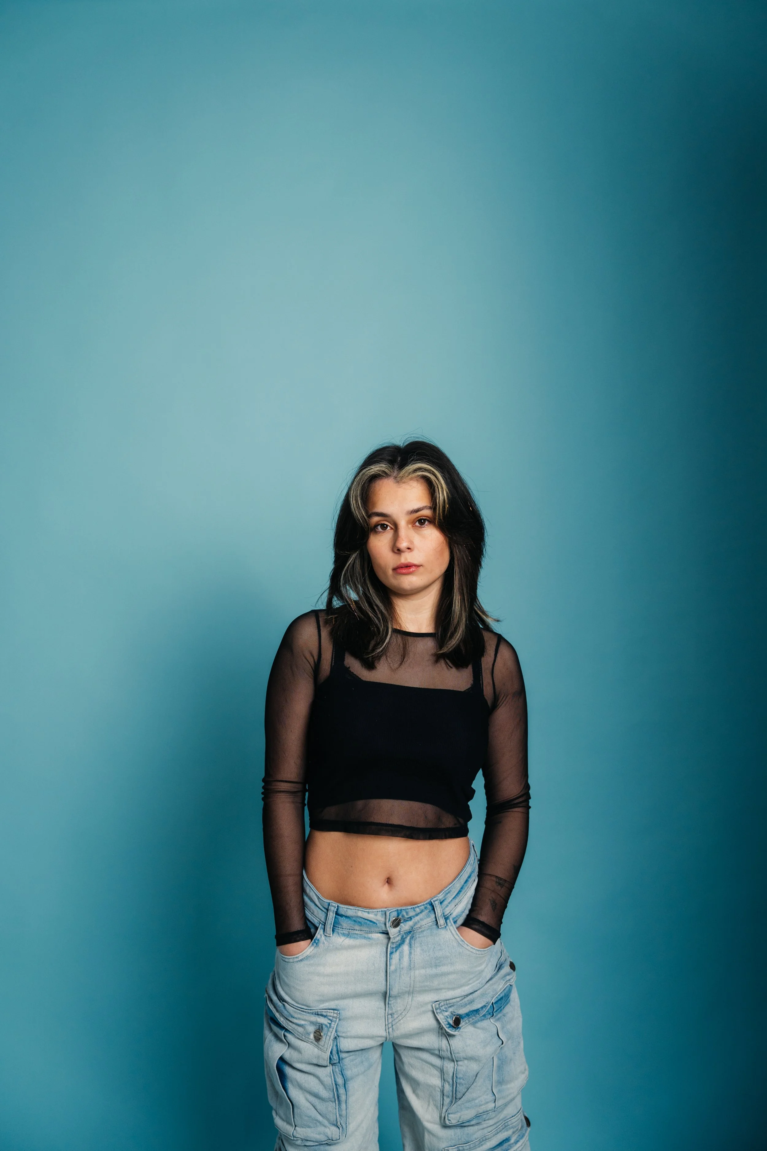 Young woman with black and blonde hair, wearing a black sheer long-sleeve top and light denim cargo pants, standing against a teal background with hands in her pockets.