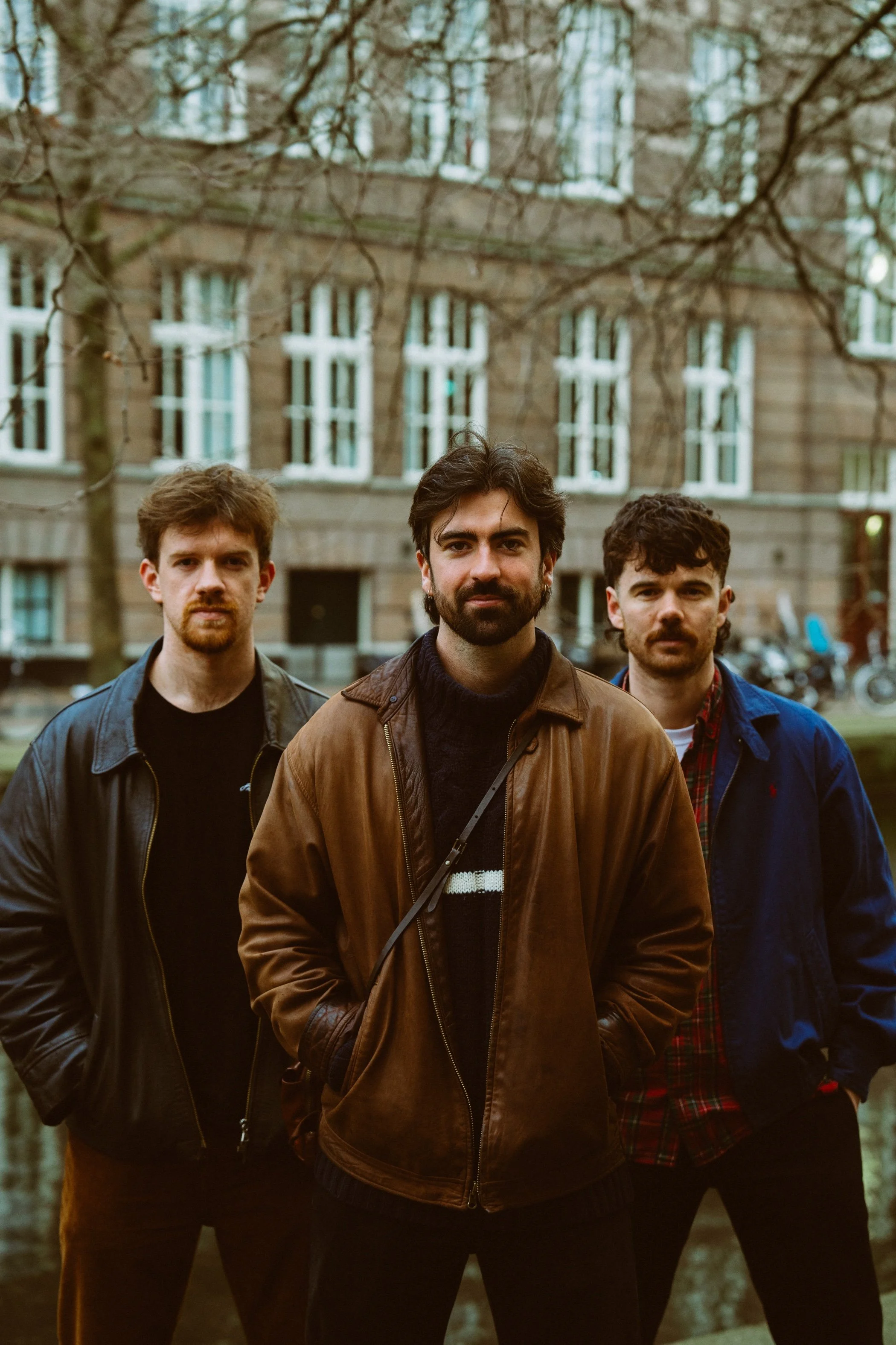 Three young men standing outdoors in front of a building with multiple windows, wearing jackets and casual clothing, with trees and bicycles in the background.