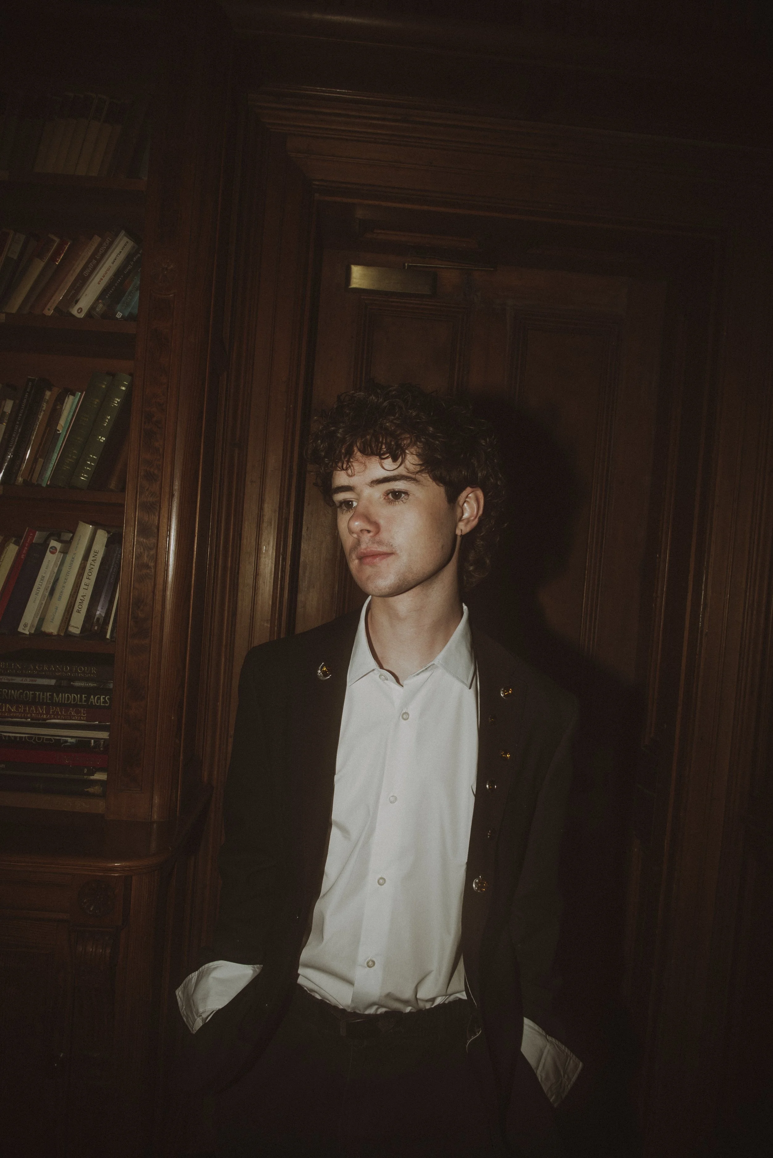 Young man with curly hair and light skin, wearing a white shirt and black blazer, standing in a wooden-paneled room near a bookshelf.