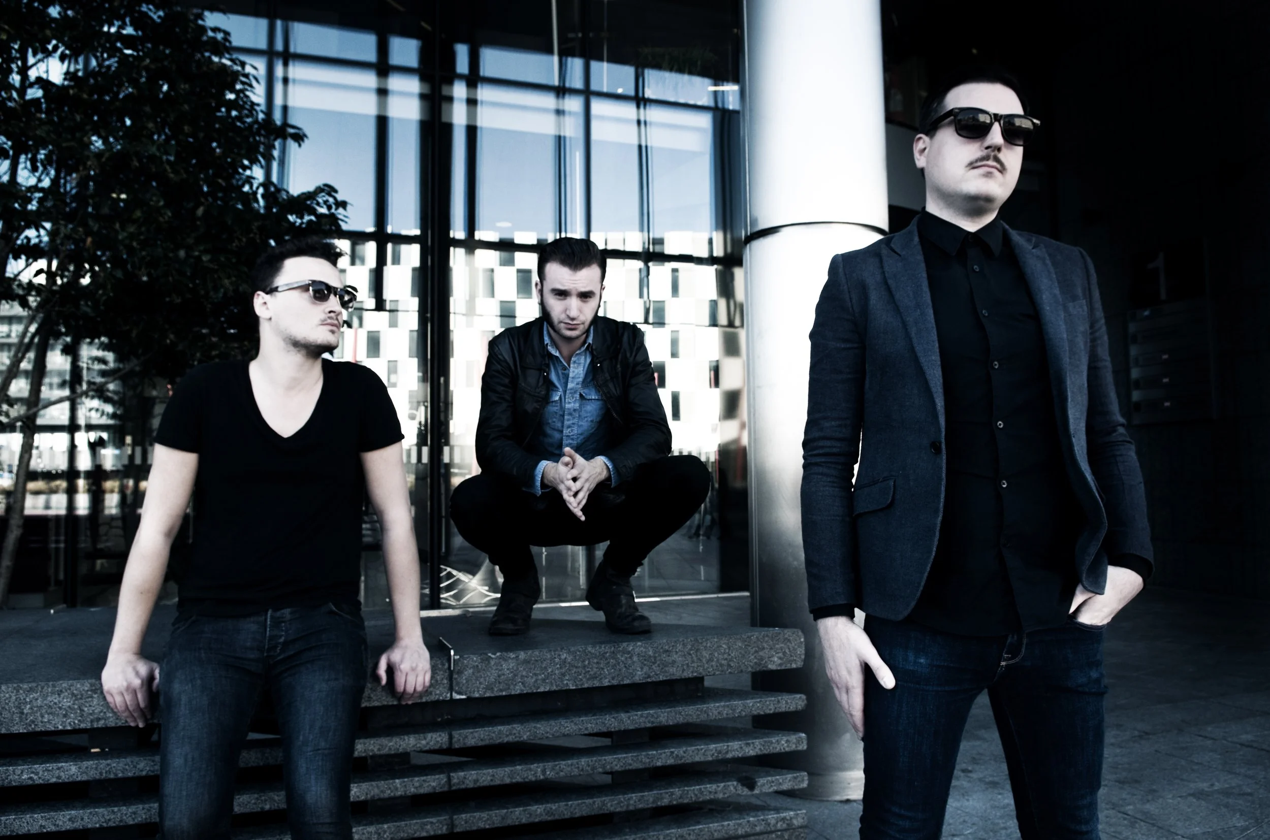 Three men with dark clothing and sunglasses outside a modern glass building, one sitting and two standing.