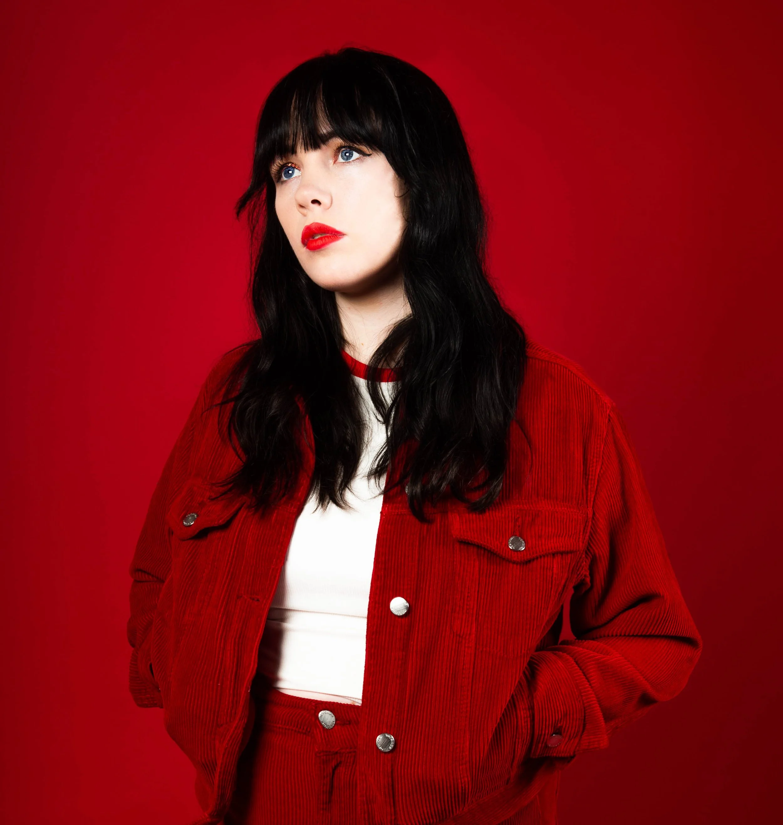 A woman with long black hair and blue eyes, wearing a red corduroy jacket over a white top, stands against a red background.