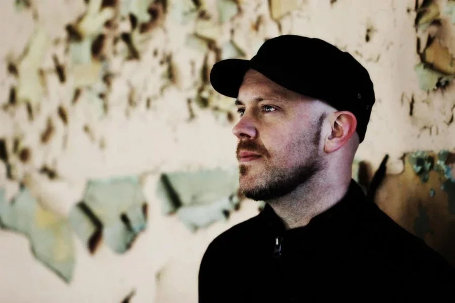 A man with a beard and mustache wearing a black cap and jacket, looking to the right, standing in front of a peeling paint wall.