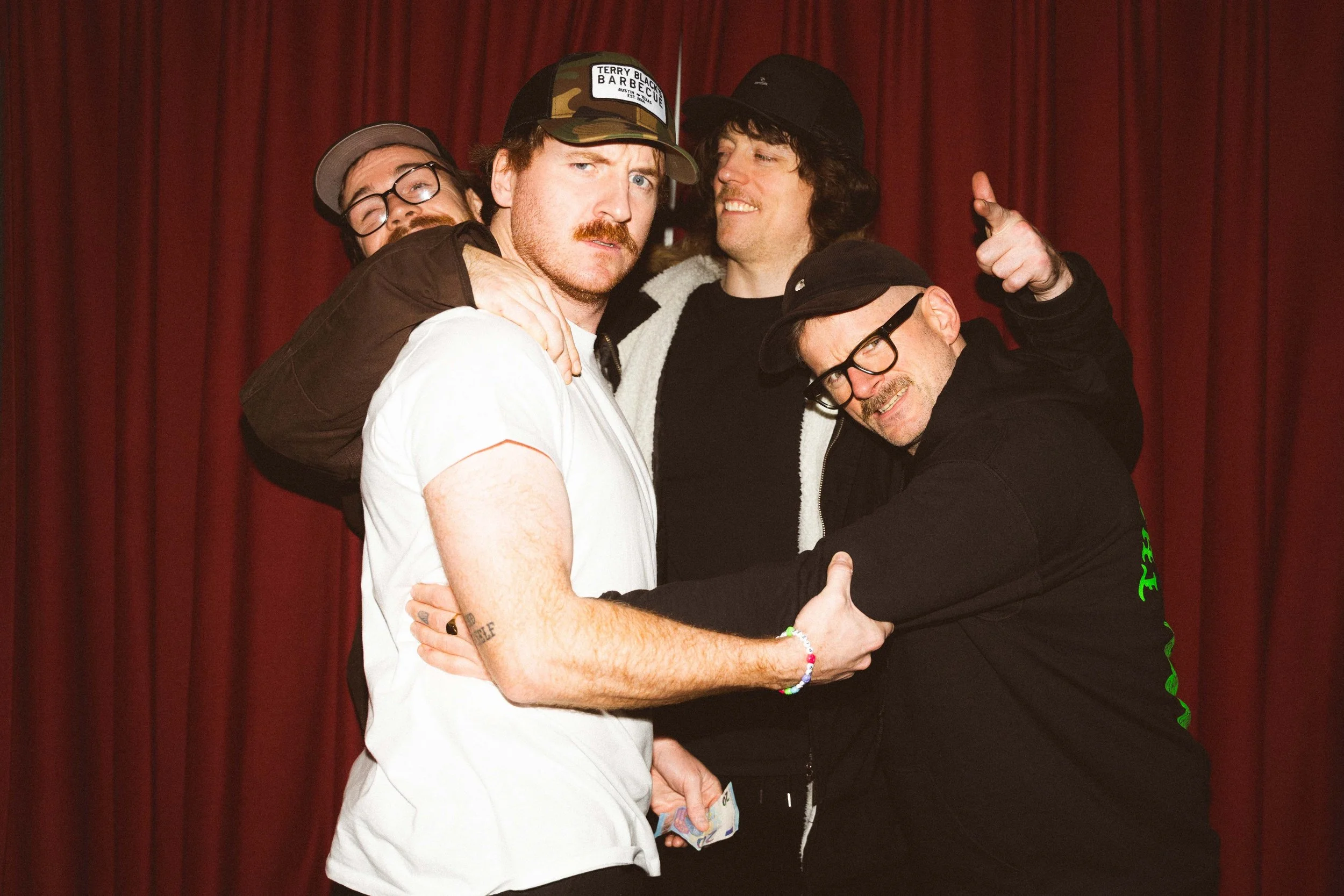 Four men posing closely together in front of a red curtain, with one man in a white shirt at the front and three men behind him, all wearing casual clothing and hats.