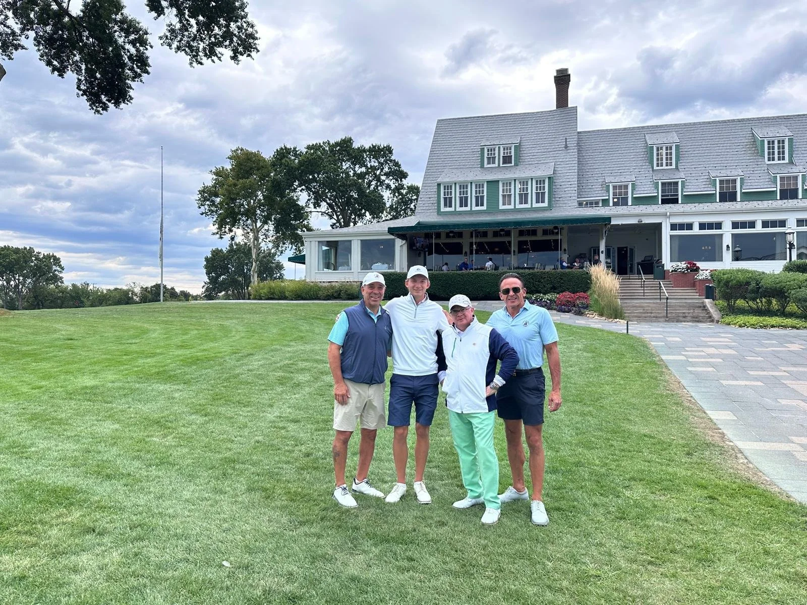 Four men standing on a golf course in front of a large clubhouse, smiling for the camera.