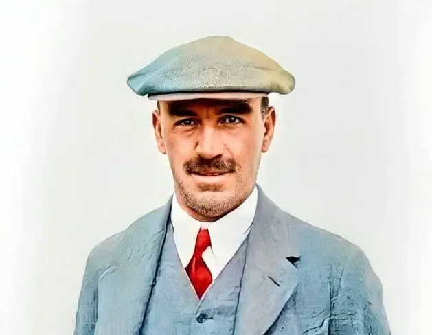 Man wearing a beige flat cap, blue suit, white shirt, and red tie, against a plain background.