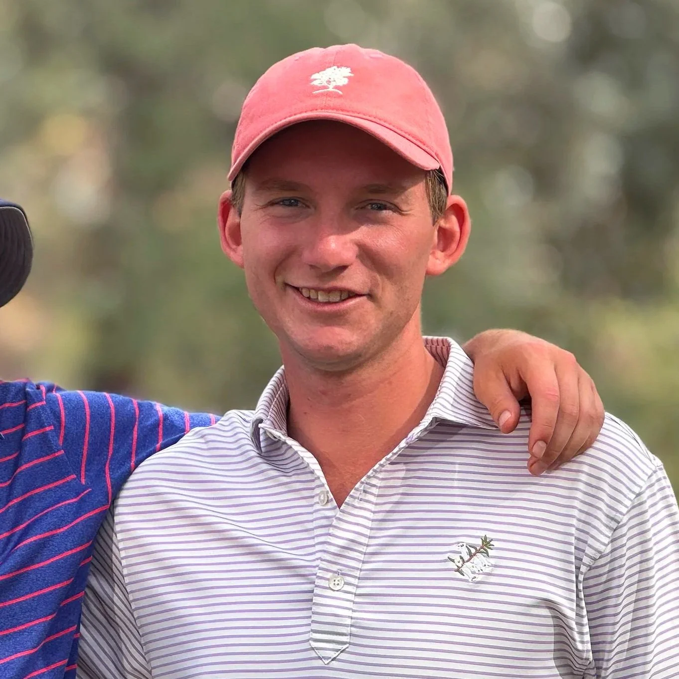 A young man smiling outdoors wearing a pink baseball cap and a striped golf shirt, with an arm around another person.