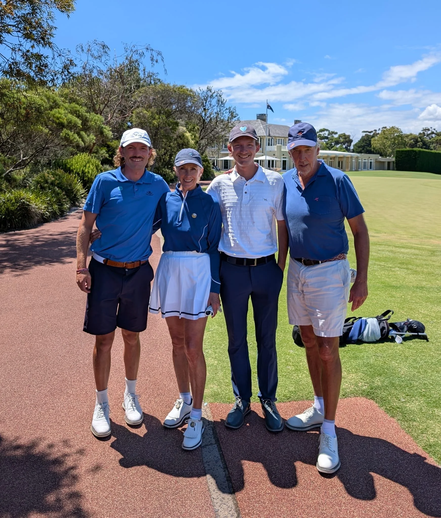Four people standing together on a golf course, smiling, with a building and trees in the background on a sunny day.