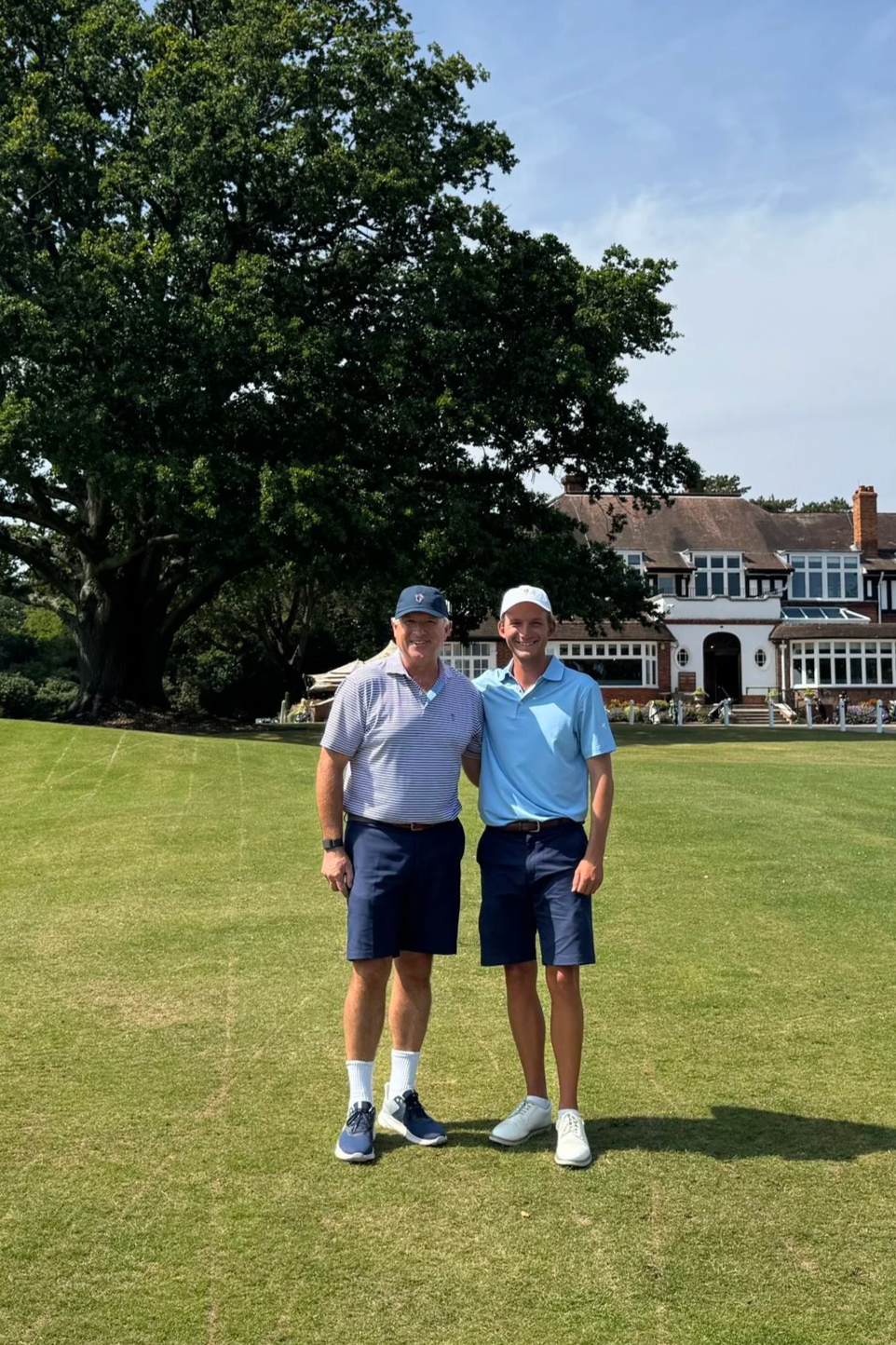 Two men standing on a golf course in front of a large house, smiling, wearing golf clothes and caps, with a large tree behind them.